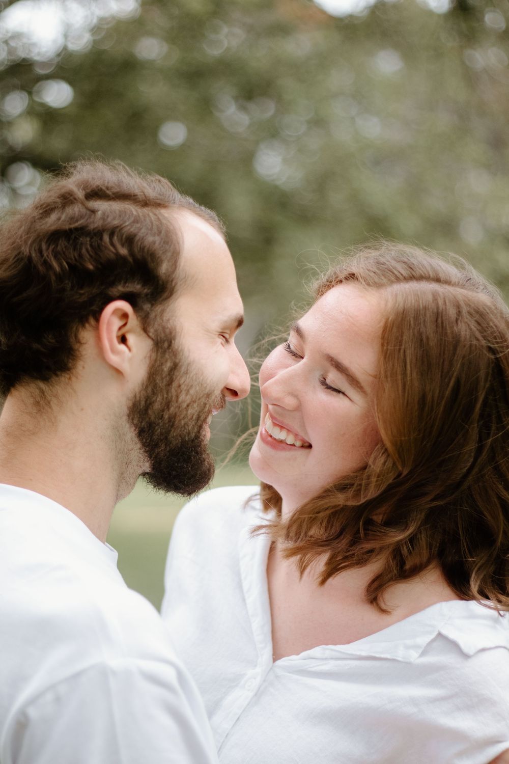 A smiling couple in white shirts share a romantic moment outdoors, nearly touching noses with trees in the background.
