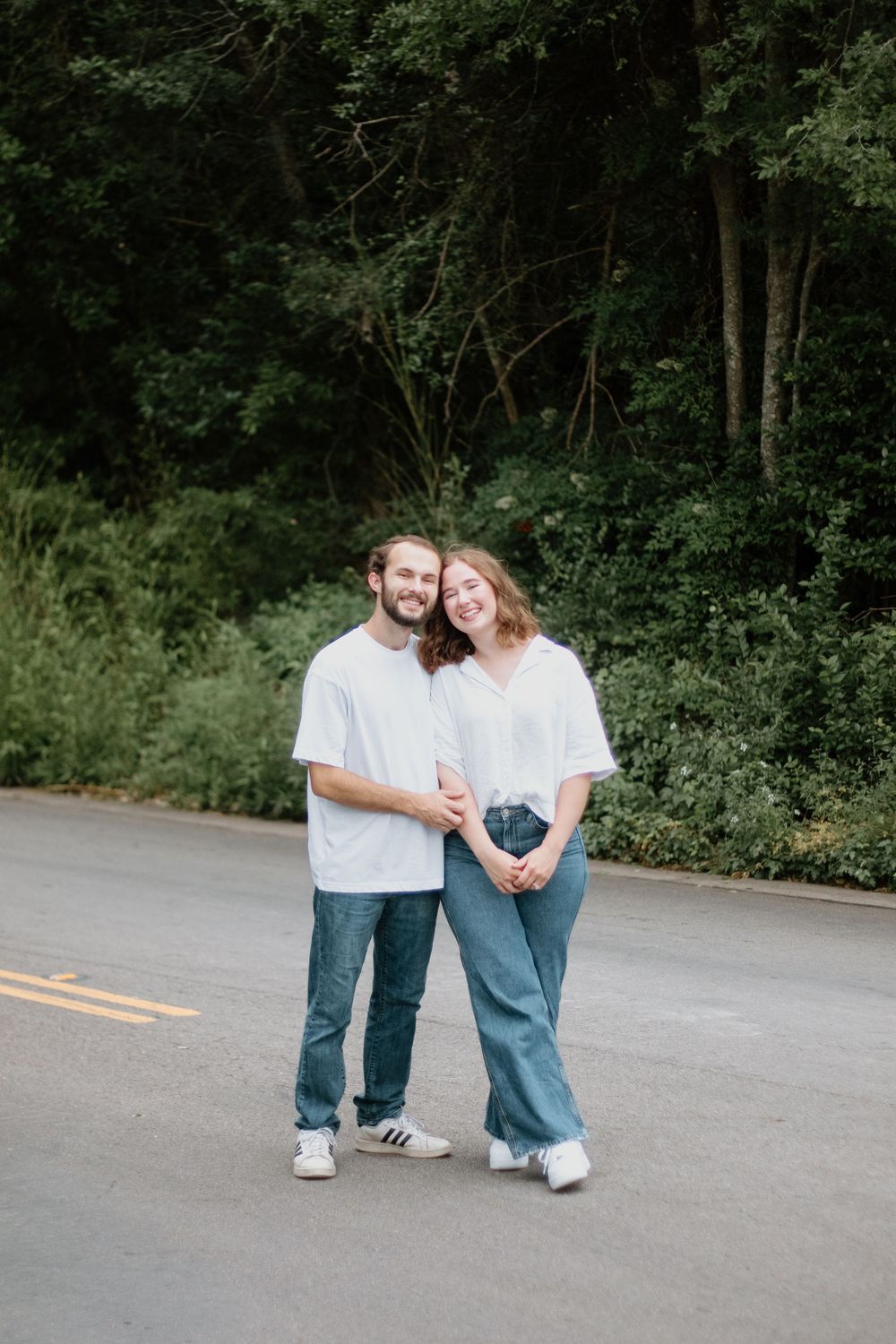 A smiling couple in white shirts and jeans poses together on a road surrounded by lush green trees.