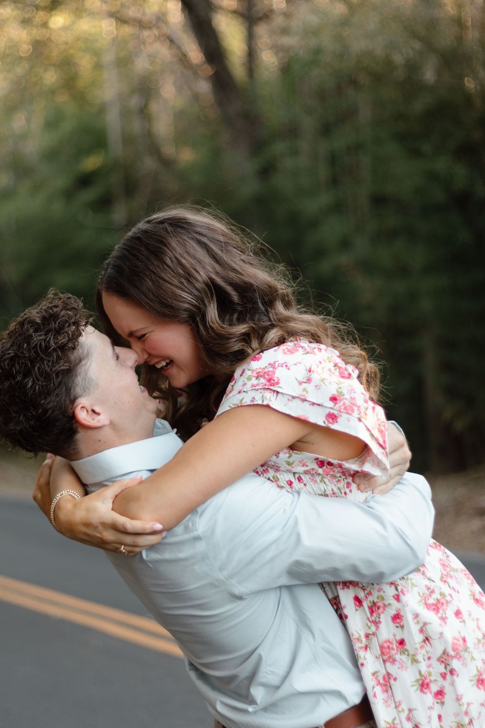 A joyful couple shares a romantic embrace on a tree-lined road, woman in floral dress lifted by her partner.