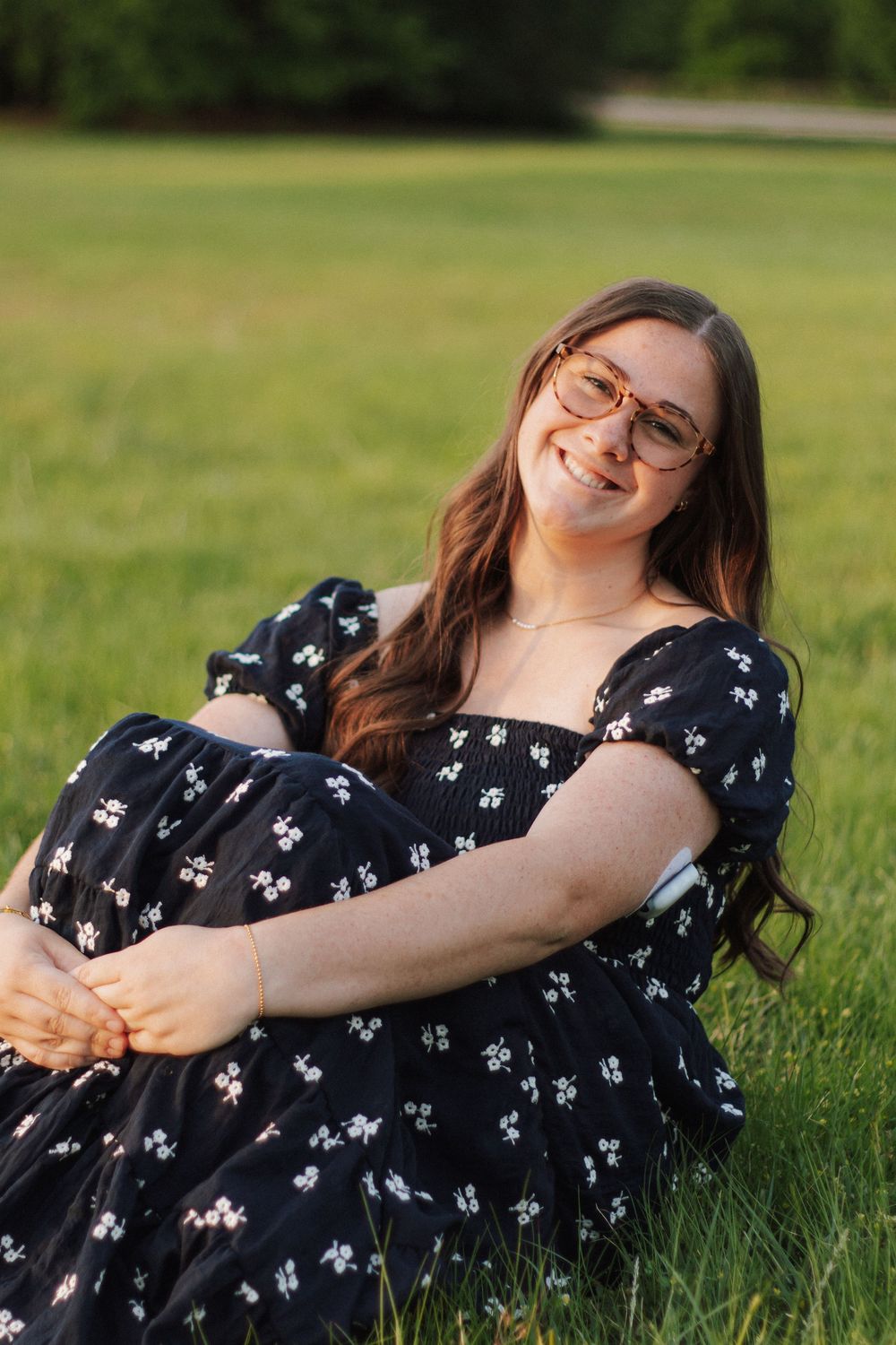 Smiling young woman with glasses in floral black dress sitting on green grass outdoors in warm sunlight.