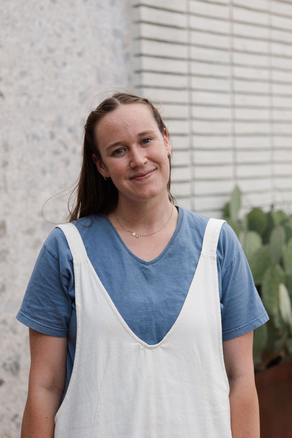 Young woman smiling outdoors in blue t-shirt and white pinafore dress, standing near a cactus plant.