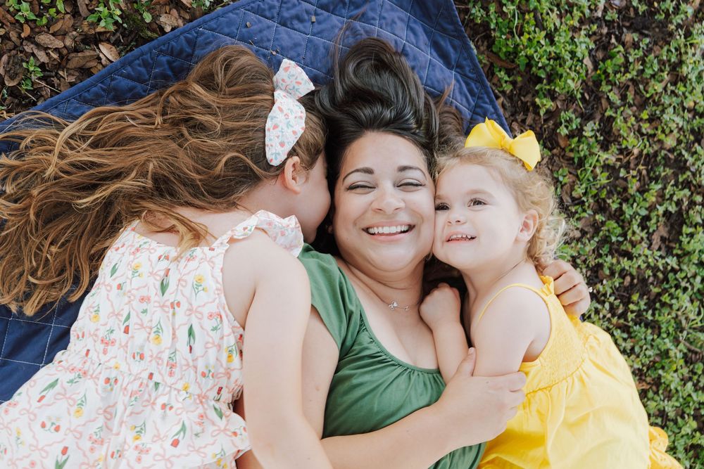 A smiling mother lies on a blanket outdoors while two young daughters in colorful dresses cuddle and kiss her cheeks.