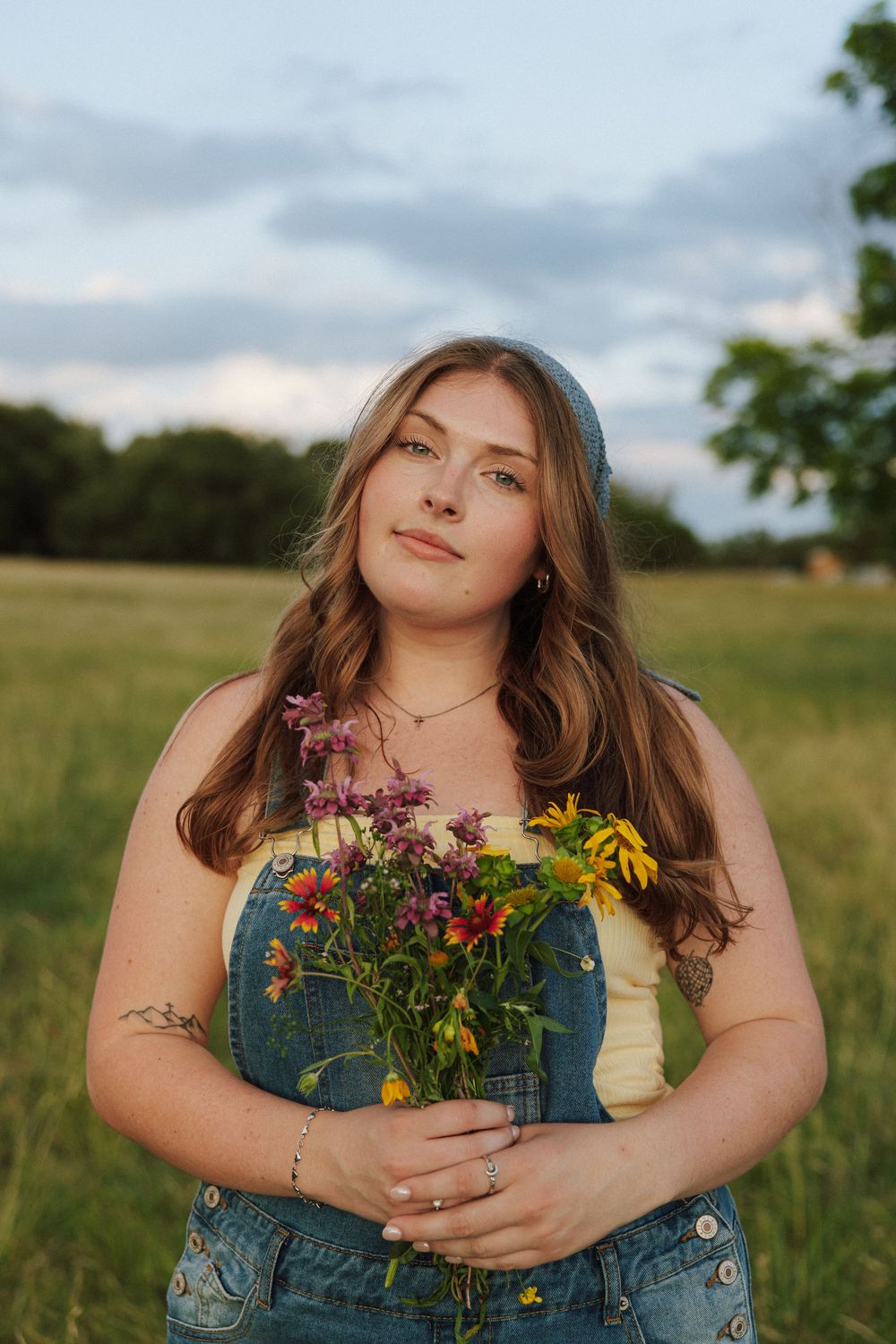 Young woman in denim overalls holding colorful wildflowers in a green field at golden hour.