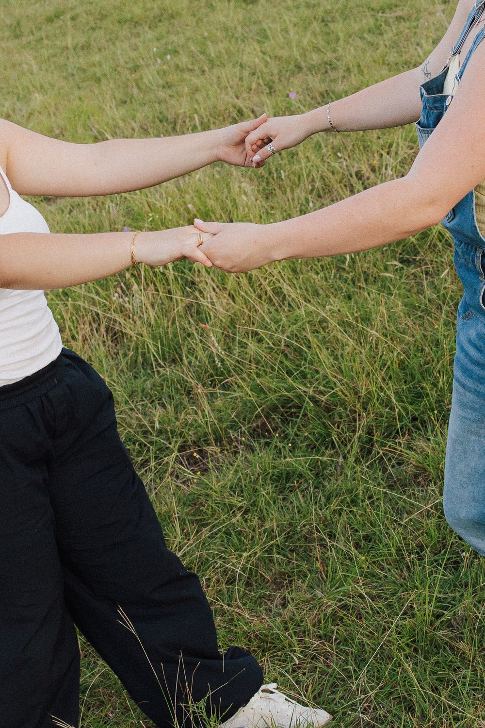 Two people holding hands in a green grassy field, one wearing black pants and white top, other in blue overalls.