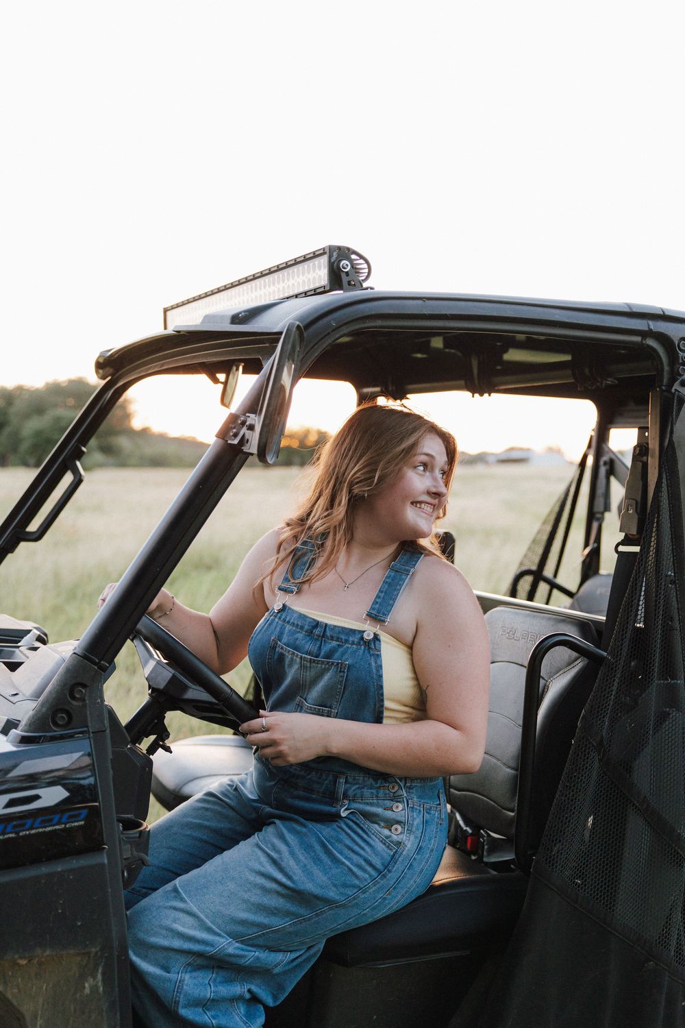 Young woman in denim overalls smiling while sitting in an off-road UTV vehicle in a scenic field at sunset.
