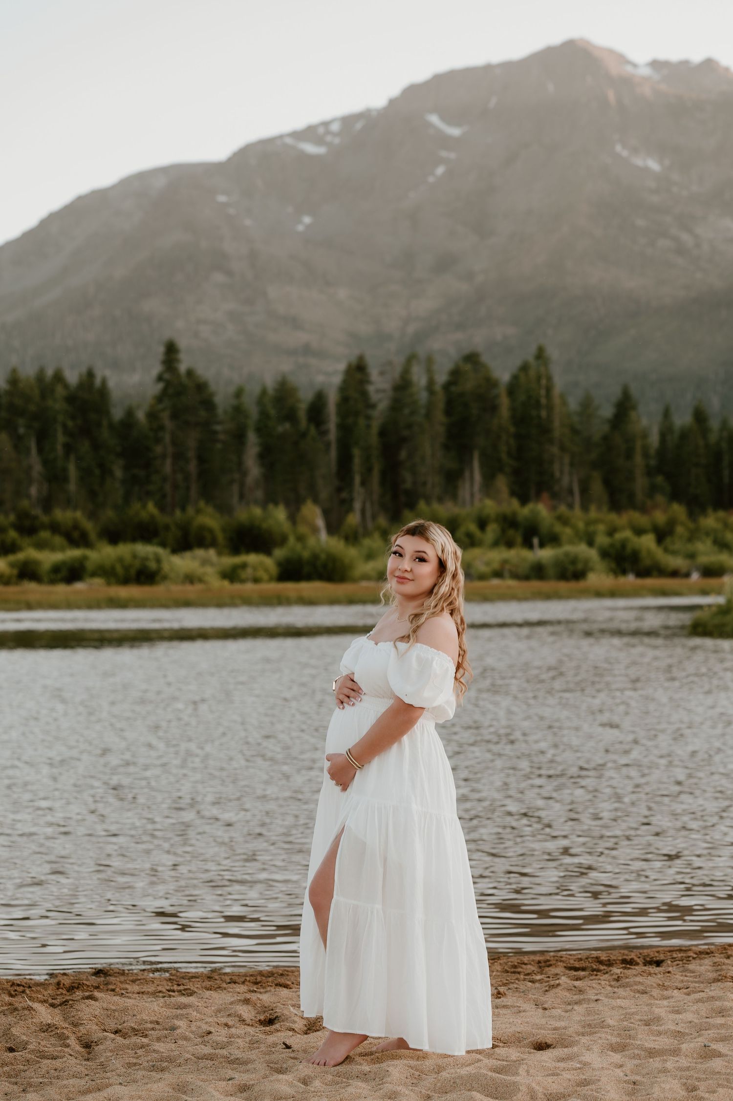 Pregnant woman in white off-shoulder maxi dress standing on sandy beach with mountain lake and pine trees backdrop.