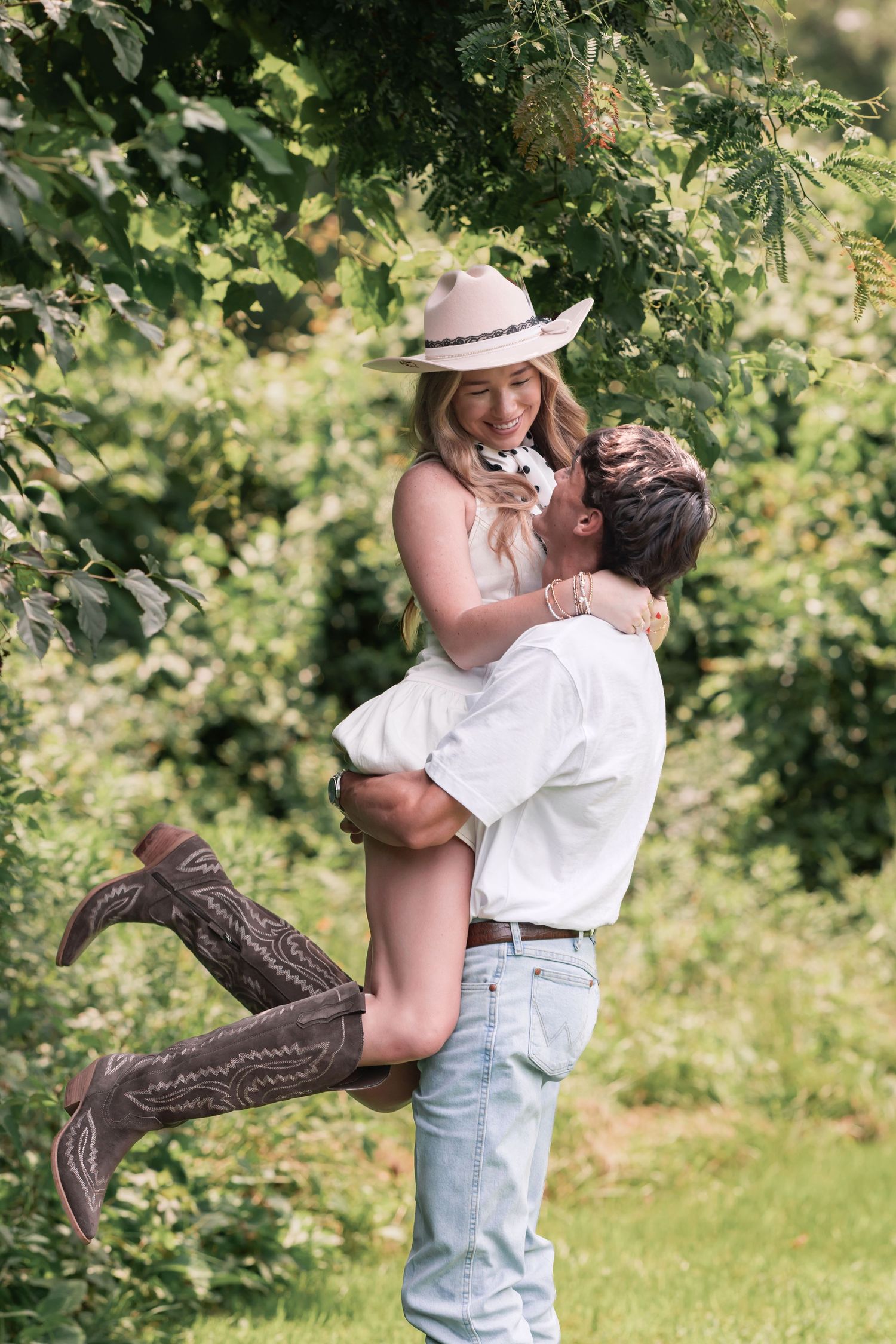 Man lifting woman in cowboy hat and boots while holding a puppy outdoors in a lush green garden.