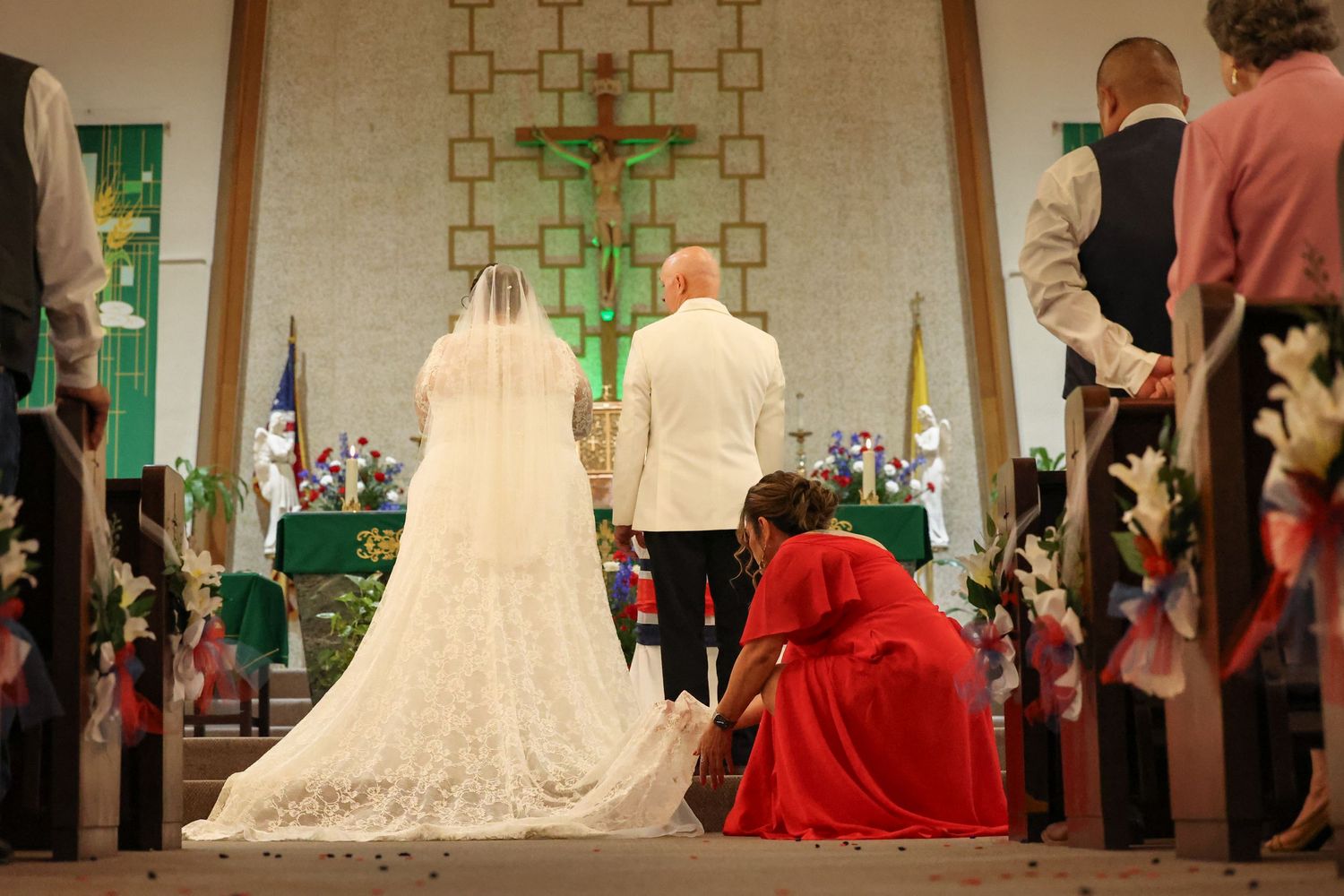 Bride and groom kneeling at church altar during wedding ceremony with attendant adjusting bridal gown train.