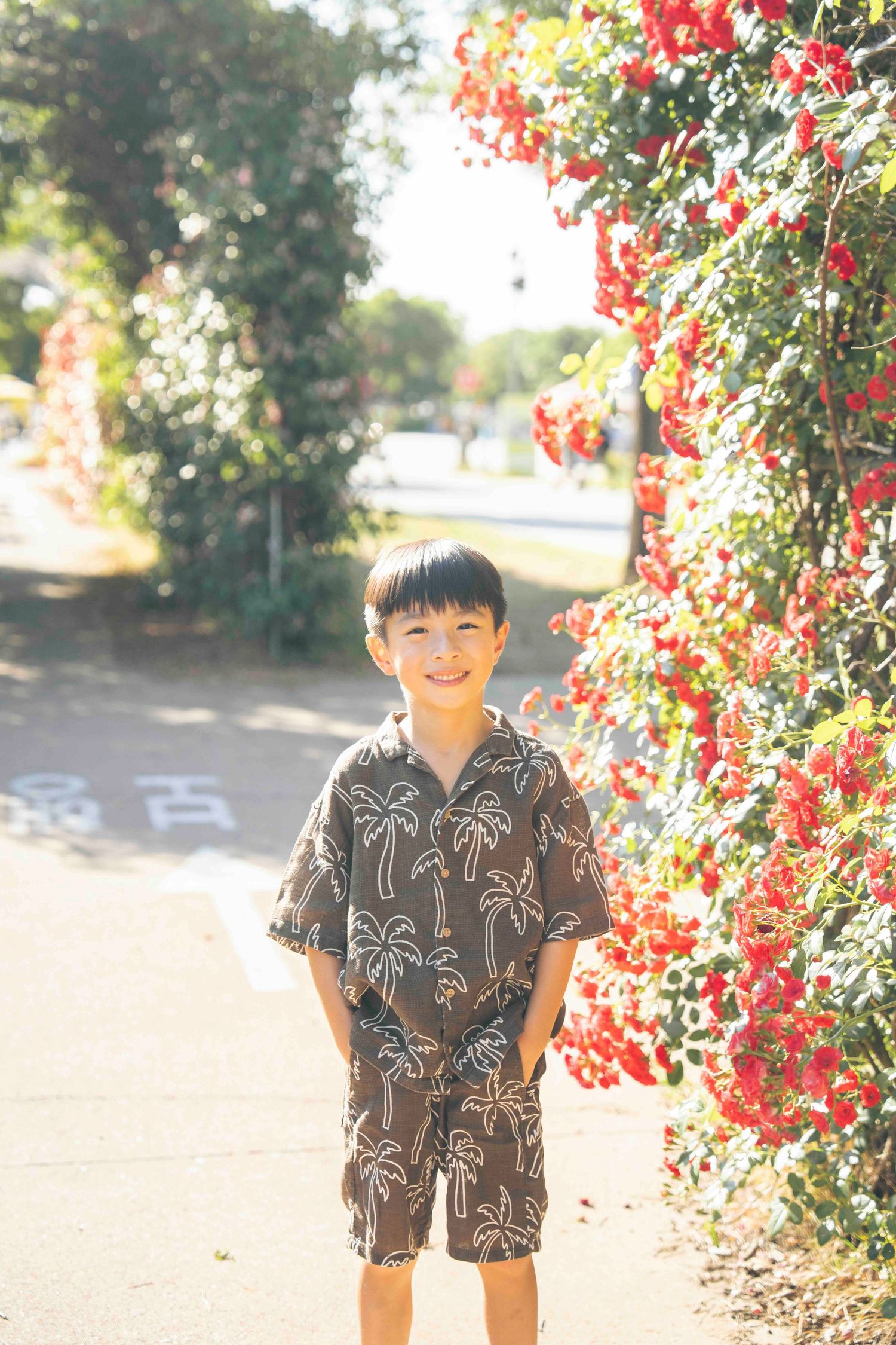 Smiling young boy in palm tree print outfit standing on sunny path beside vibrant red flowering bougainvillea.
