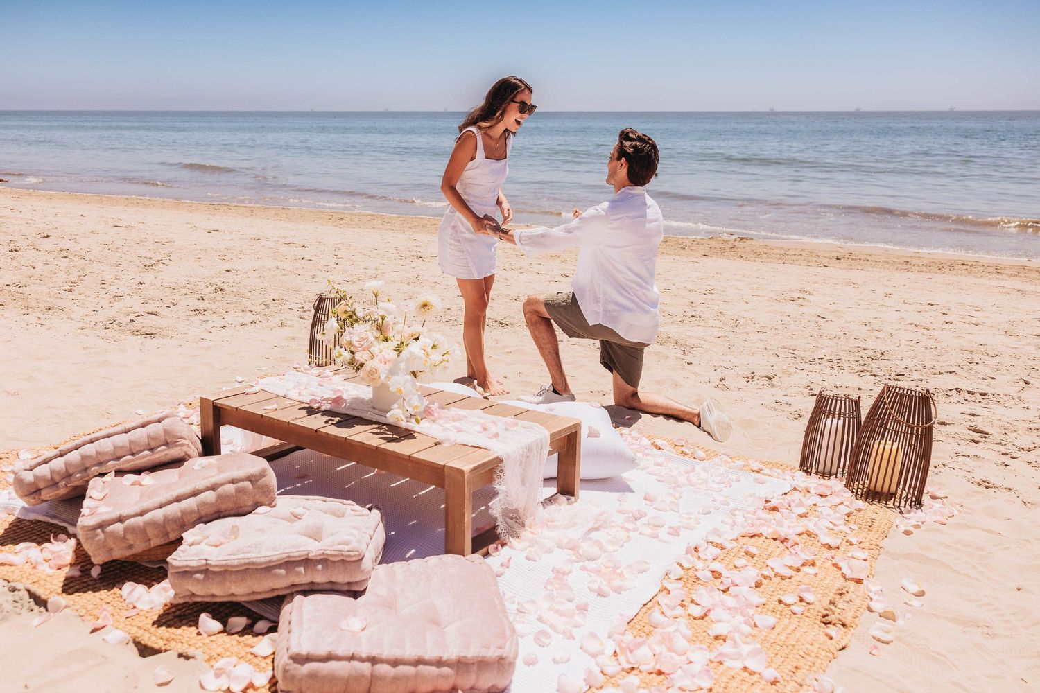 Romantic beach proposal with man on one knee, rose petals, cushions, and ocean backdrop on sunny day.