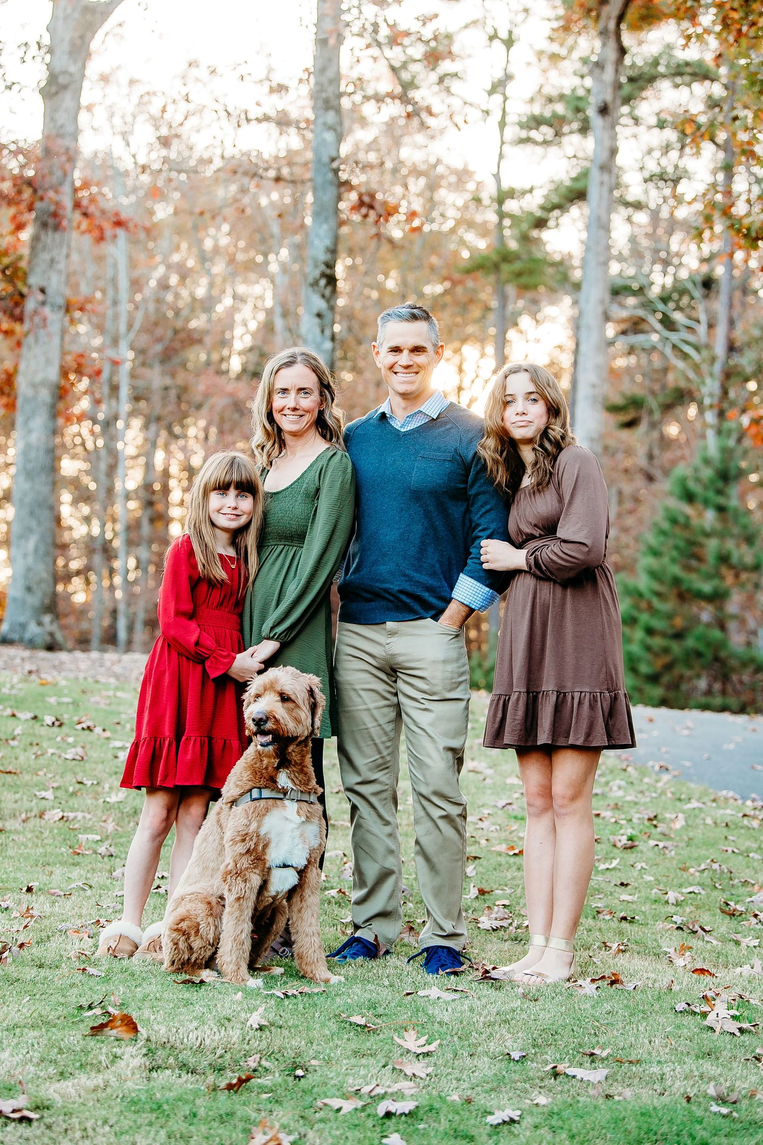 Family of four with dog posing outdoors in fall foliage, wearing coordinated outfits in red, green, blue, and brown.