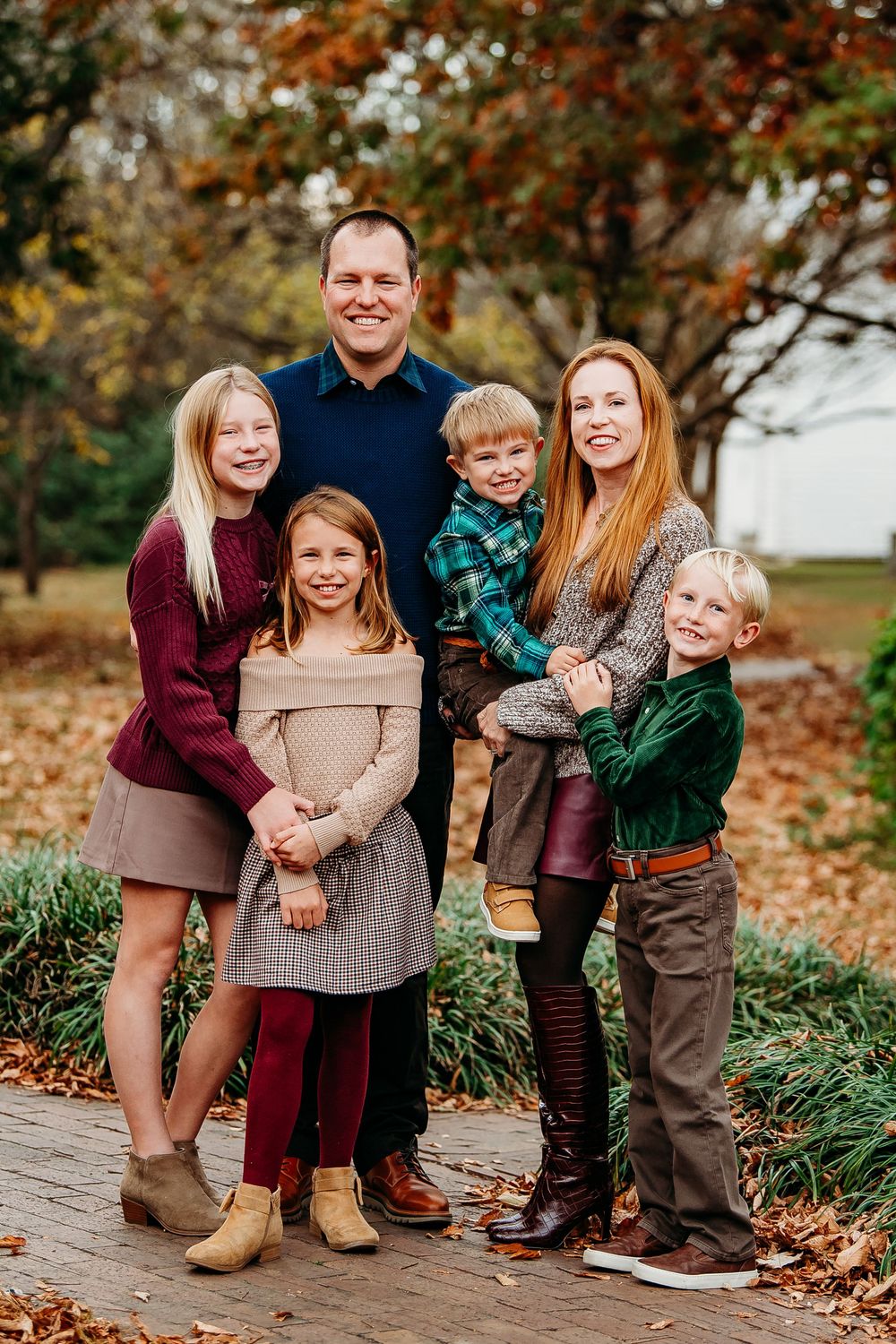 Family of six posing on a fall leaf-covered path, wearing coordinated burgundy, green, and neutral tones.