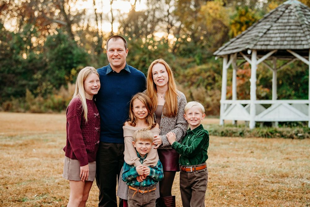 Family of five smiling together in an autumn field near a white gazebo at golden hour.