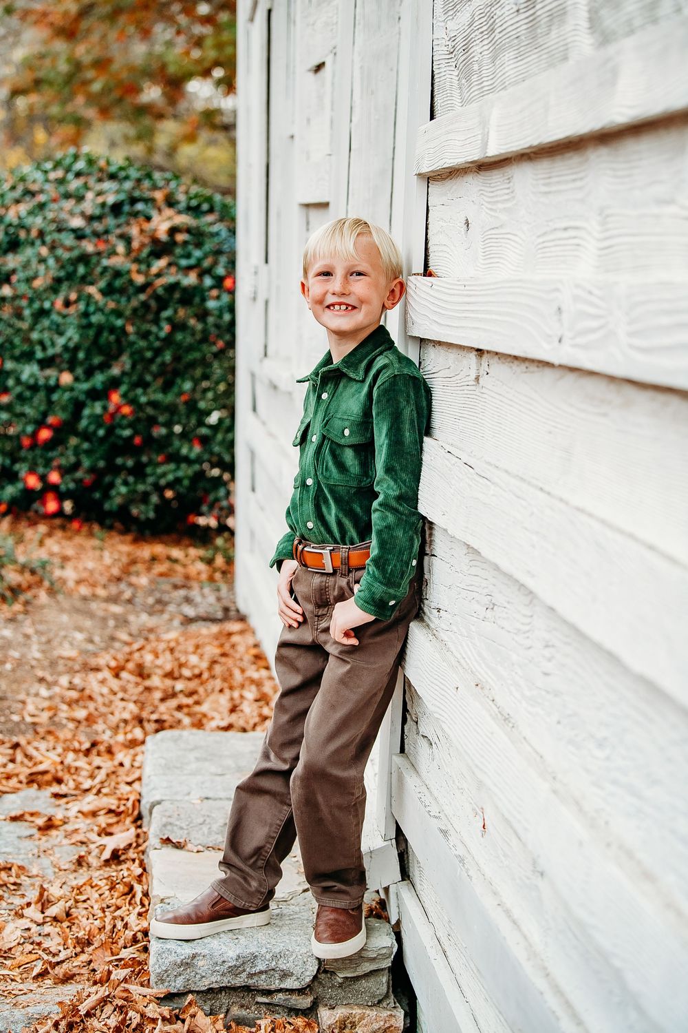 Smiling blonde boy in a green shirt and brown pants leaning against a white wooden wall in autumn.