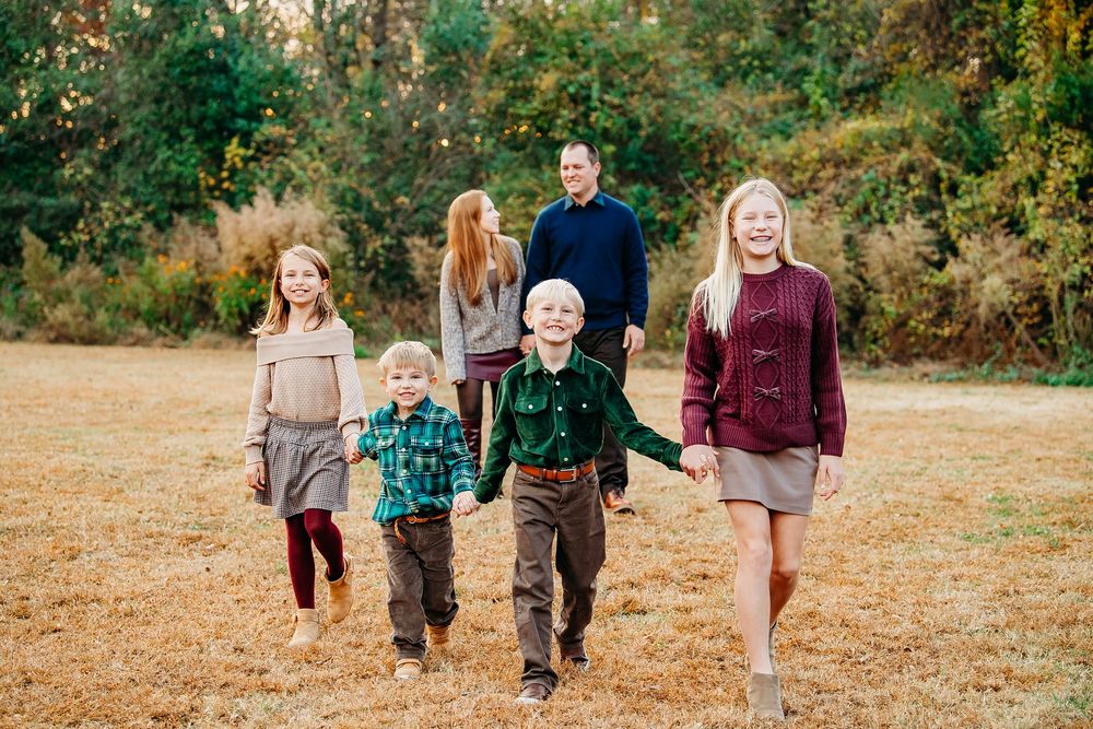 Family of six walking through an open autumn field holding hands in coordinated fall outfits.