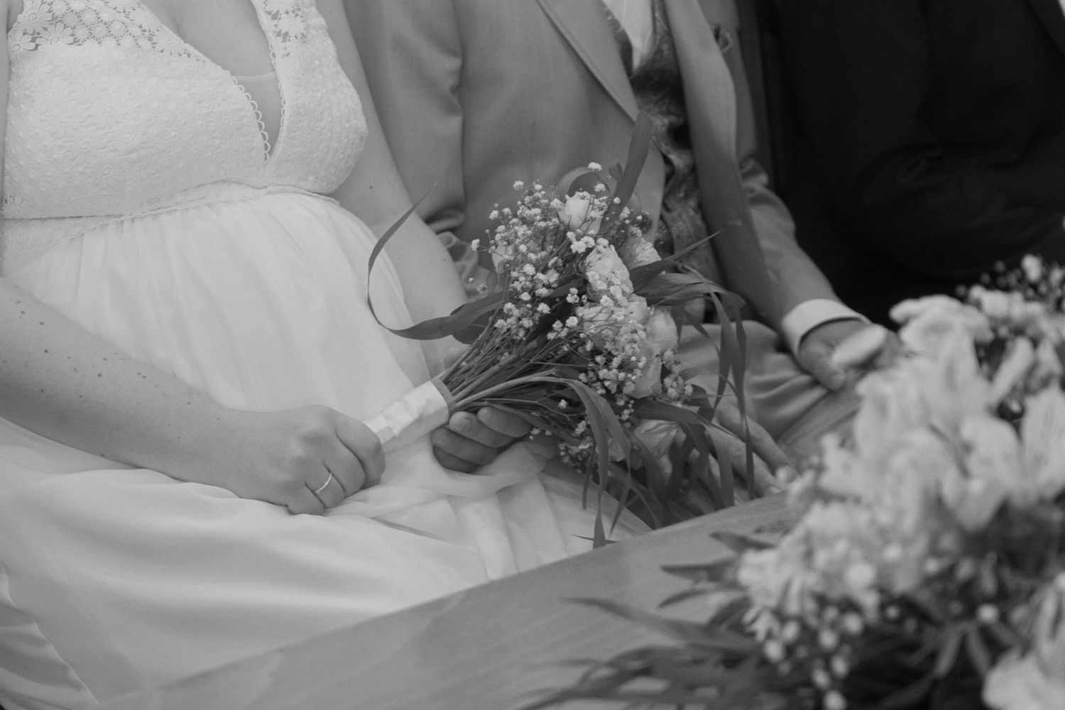 Black and white wedding photo of bride holding floral bouquet with groom seated beside her during ceremony.