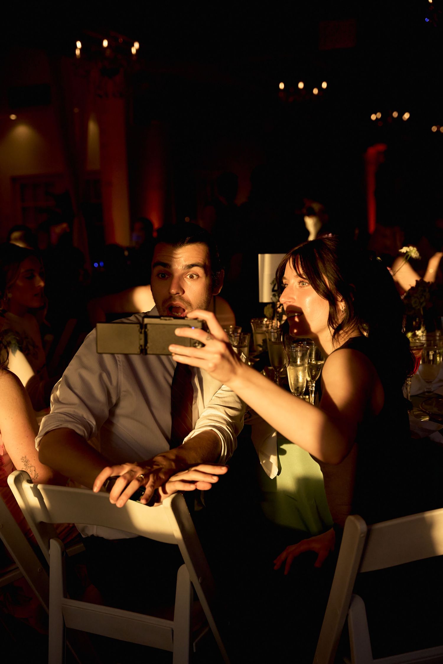 Couple taking a selfie at a dimly lit formal event, man in tie and woman in black dress at a banquet table.