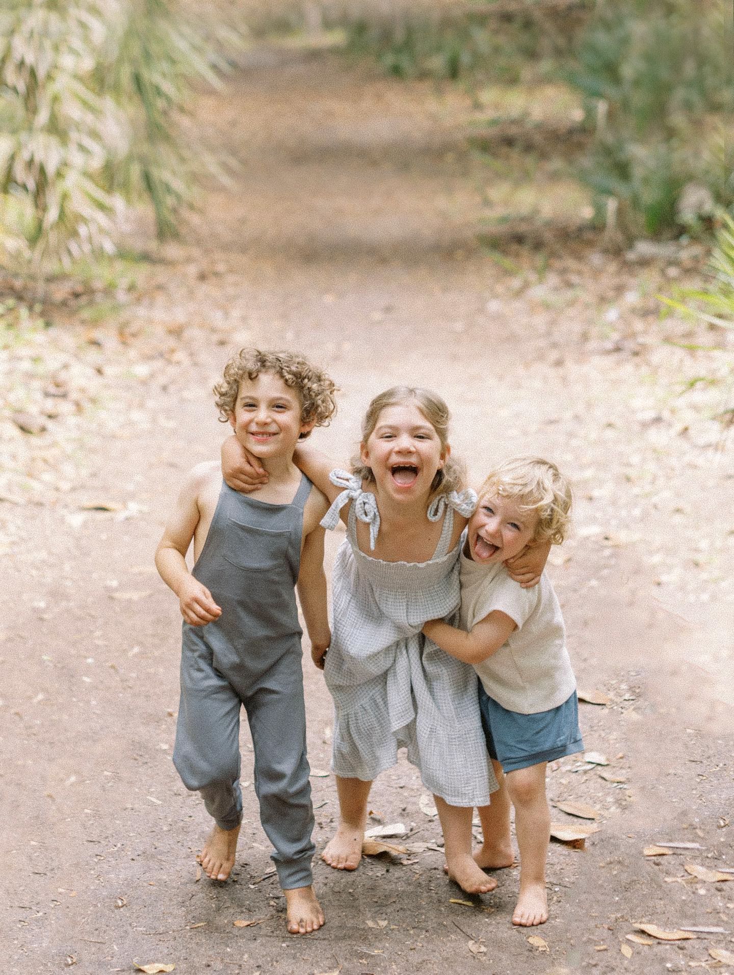 Three barefoot children laugh and hug on a dirt path surrounded by lush greenery in a candid outdoor portrait.
