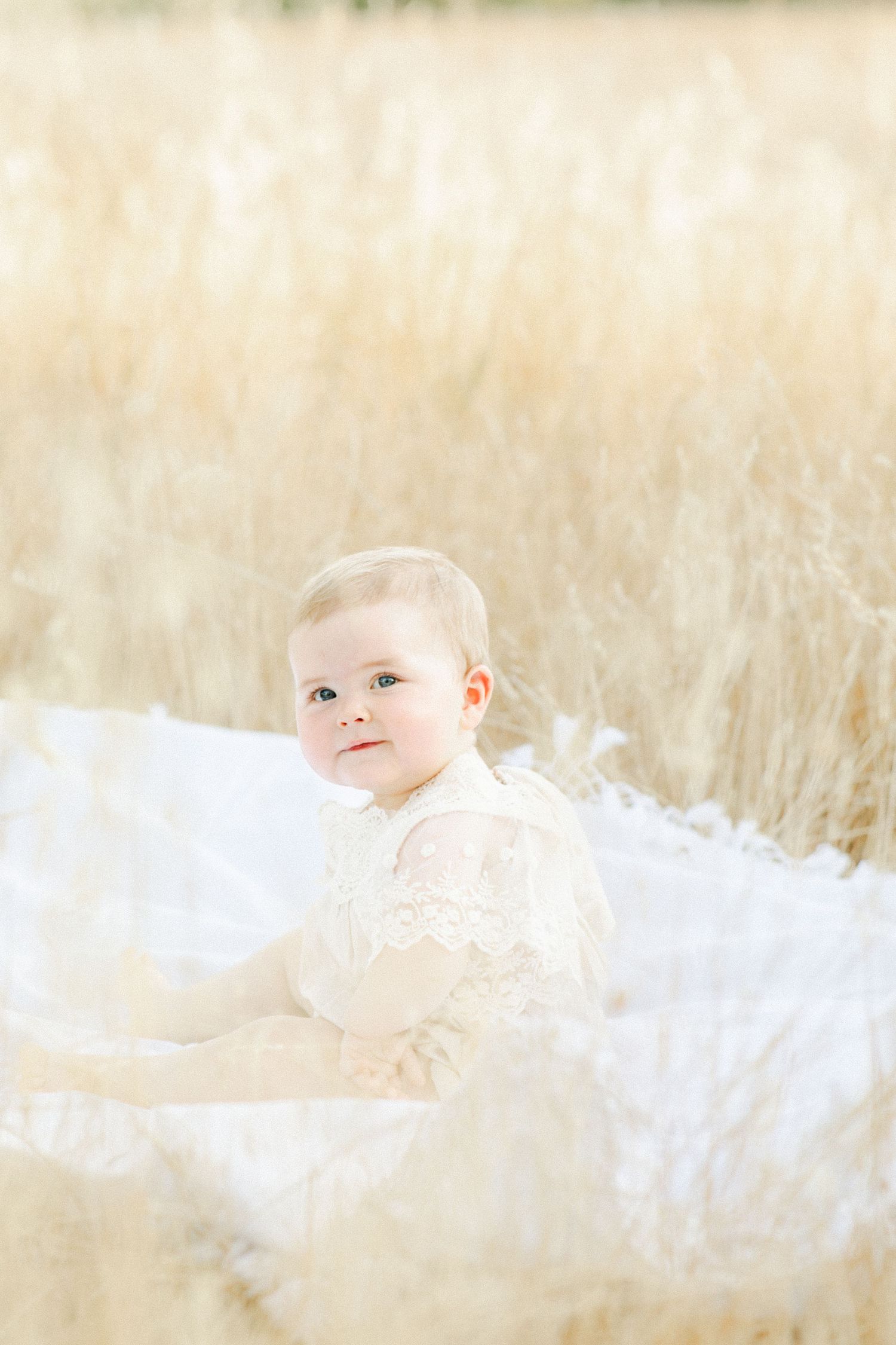 A baby in a cream lace outfit sits on a white blanket surrounded by golden wheat grass in a bright outdoor setting.