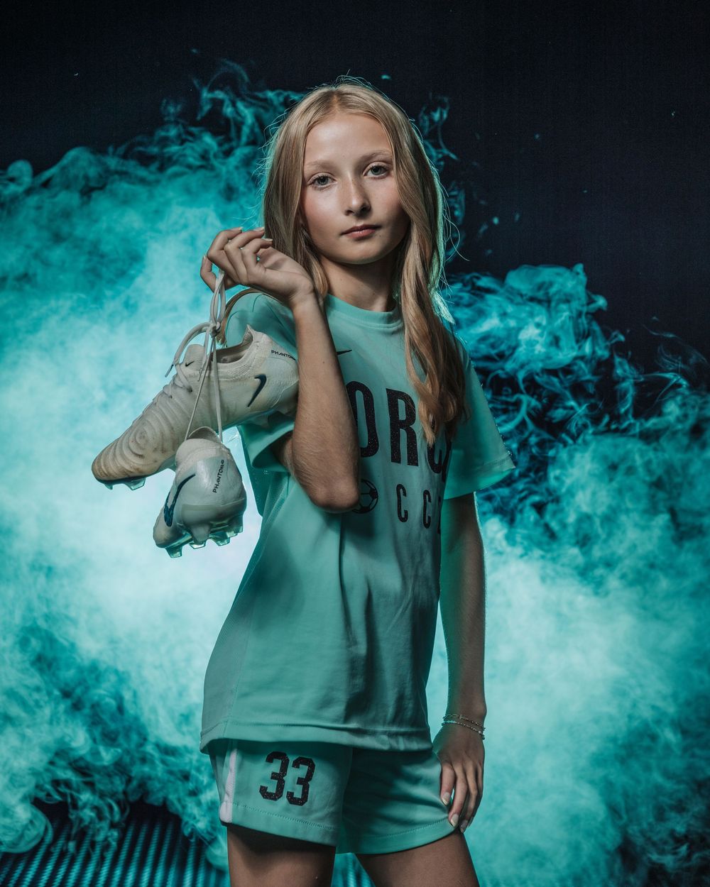 Young girl in mint green soccer uniform #33 holds cleats over shoulder against dramatic teal smoke background.