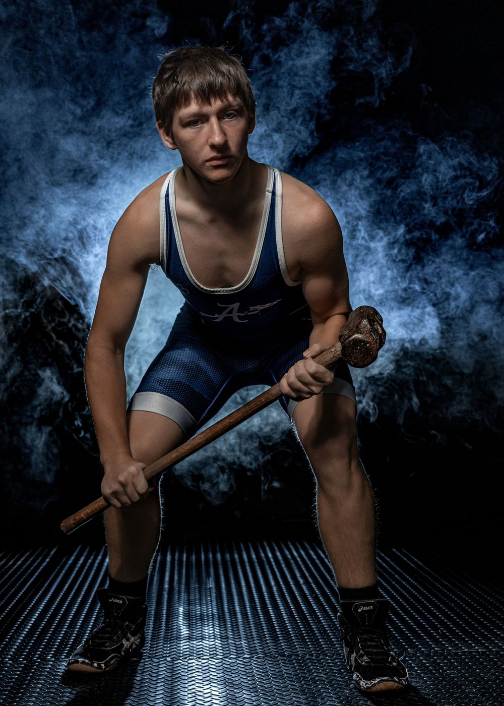 Young wrestler in blue singlet crouches dramatically holding a wooden stick on dark mat with blue smoke background.