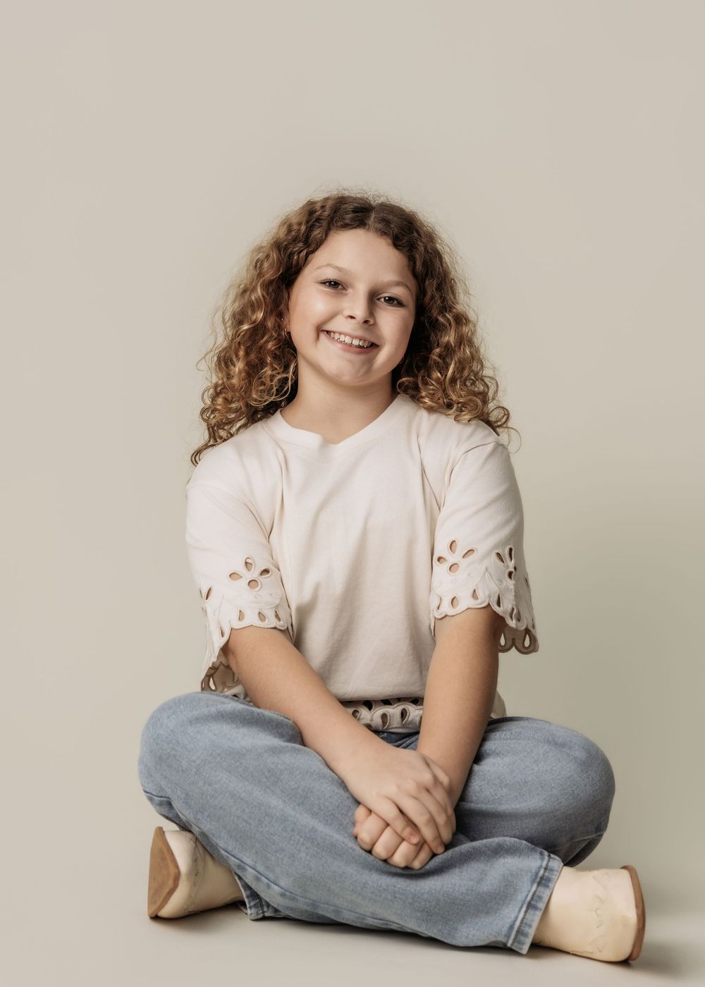 Smiling girl with curly hair sitting cross-legged, wearing a cream eyelet top and jeans, studio portrait.