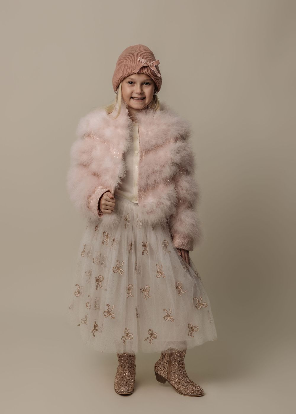 Girl in pink faux fur jacket, tulle skirt with floral embroidery, glitter boots, and pink knit hat poses gracefully.