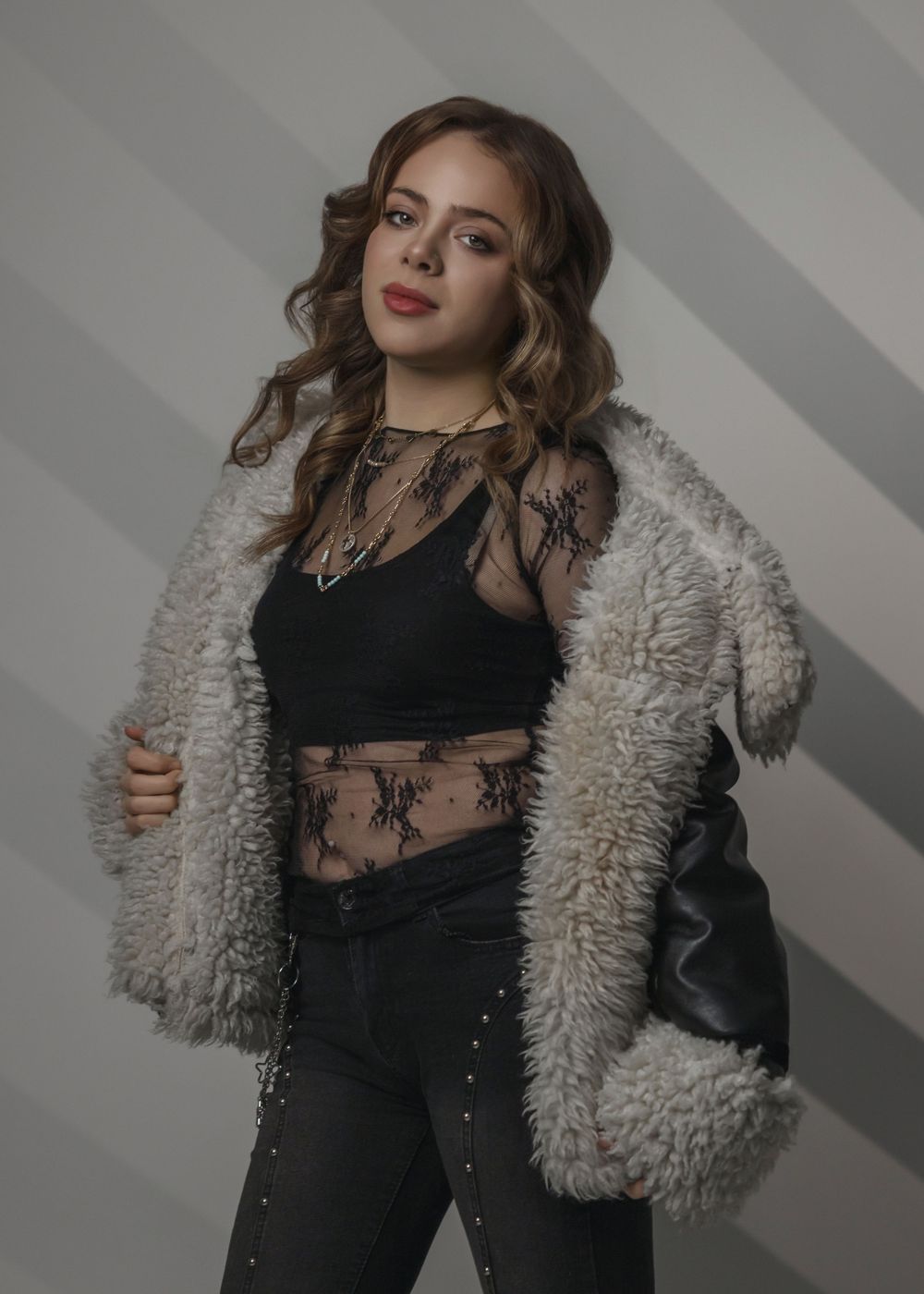 Young woman in fluffy gray coat over black crop top and dark jeans poses confidently against striped background.