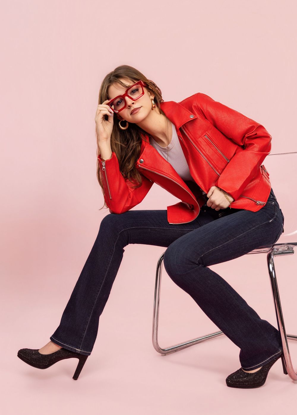 Woman in red leather jacket, dark flare jeans, black heels, and red glasses poses on chair against pink background.