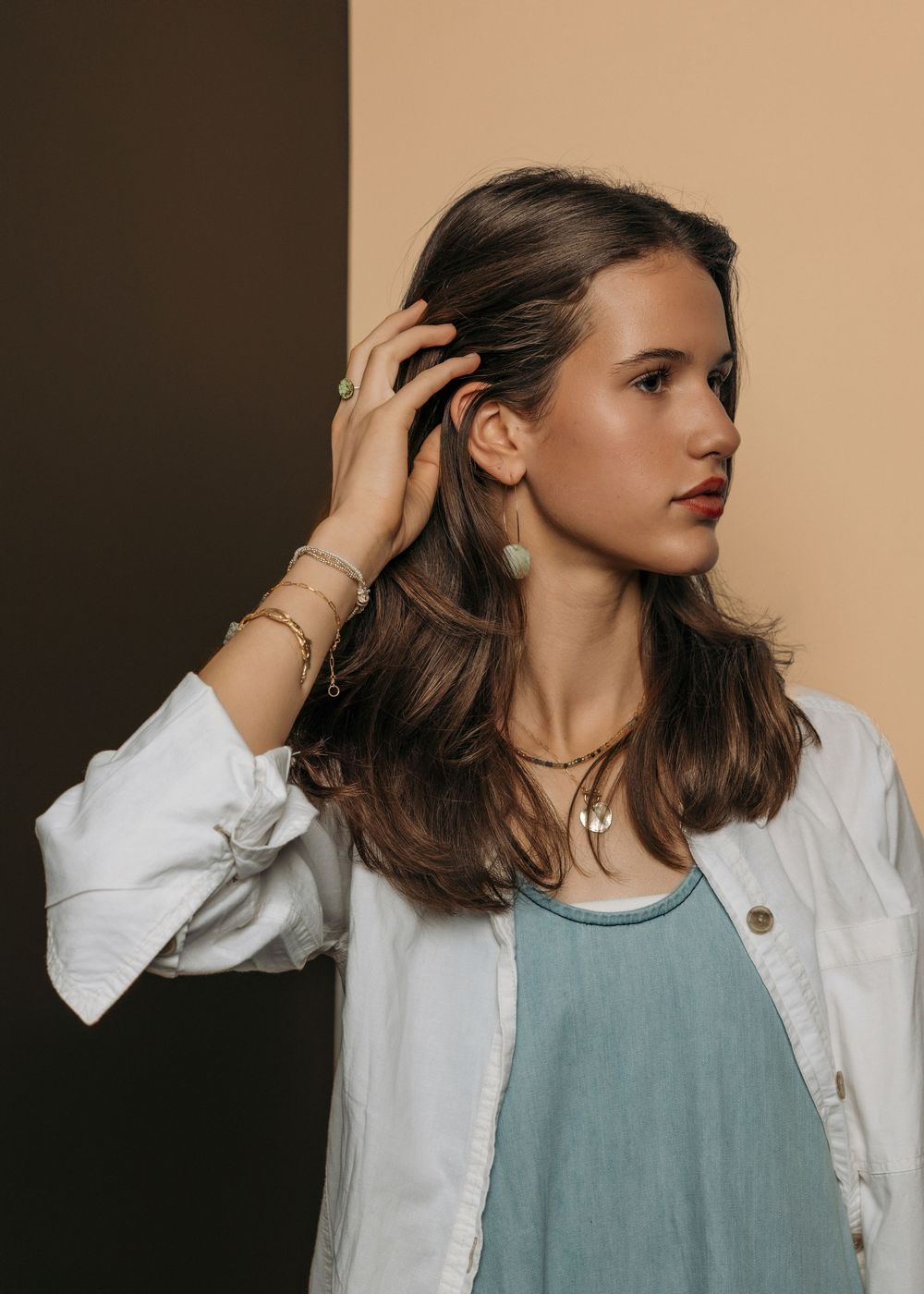 Young woman in white jacket and teal top wearing gold jewelry, posing with hand in brown hair against beige background.