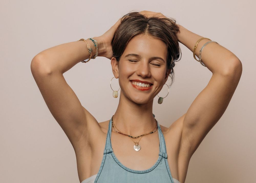 Smiling young woman with eyes closed, hands in hair, wearing blue tank top, gold earrings, and layered necklaces.