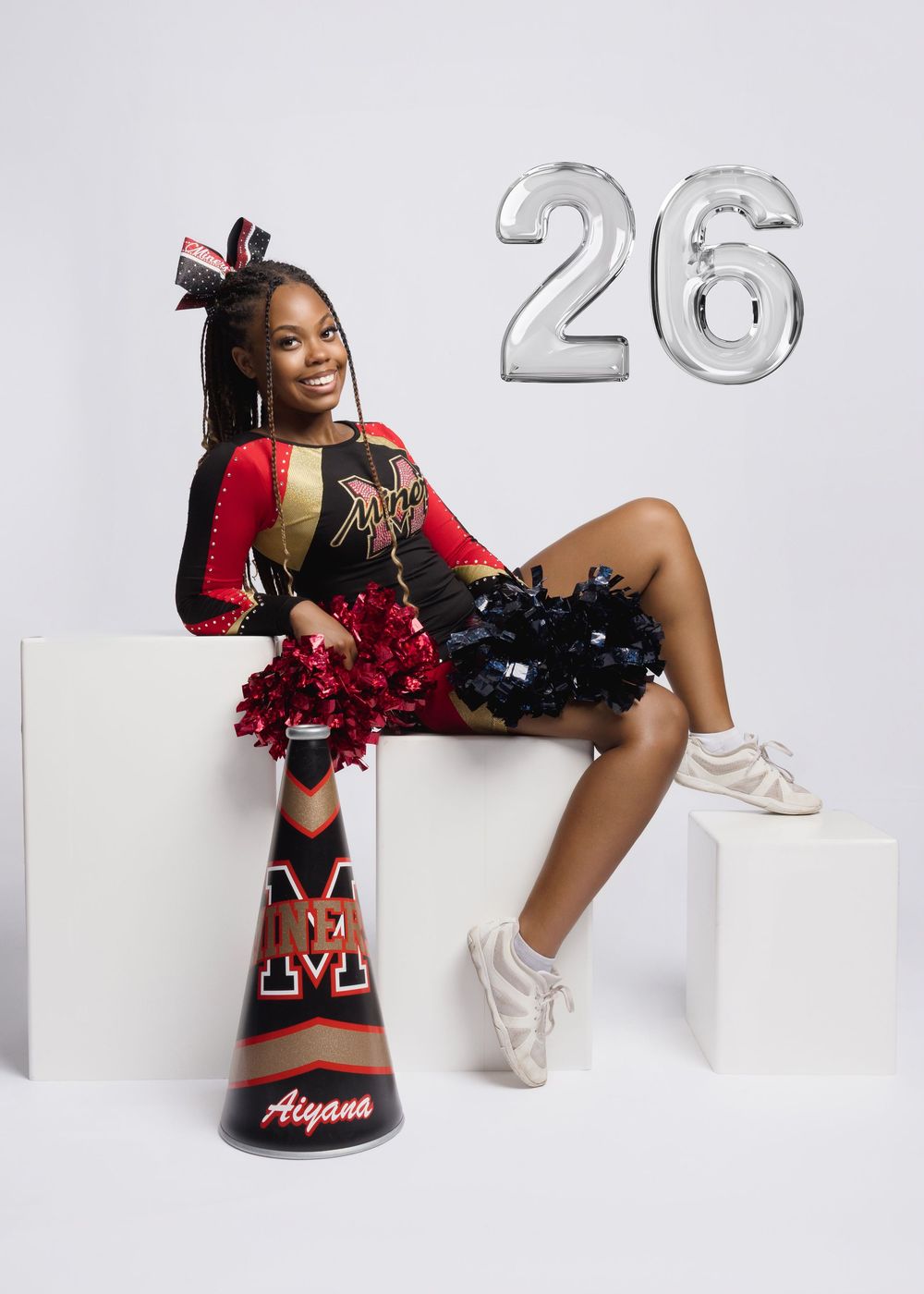 Cheerleader named Aiyana poses in red and black uniform with pom-poms beside number 26 balloons.