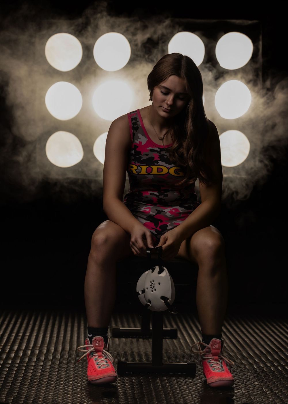 Female athlete in pink camo uniform sits on bench holding soccer ball under dramatic studio lights with smoke effects.