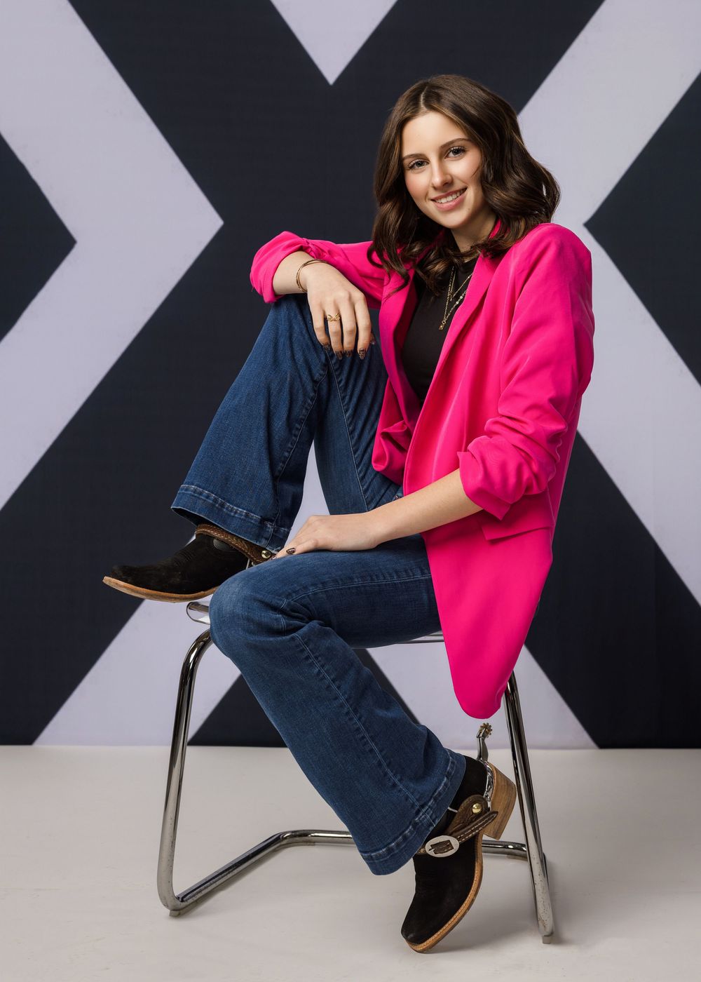 Woman in pink blazer and jeans sits on chrome chair, smiling, against geometric black and white background.