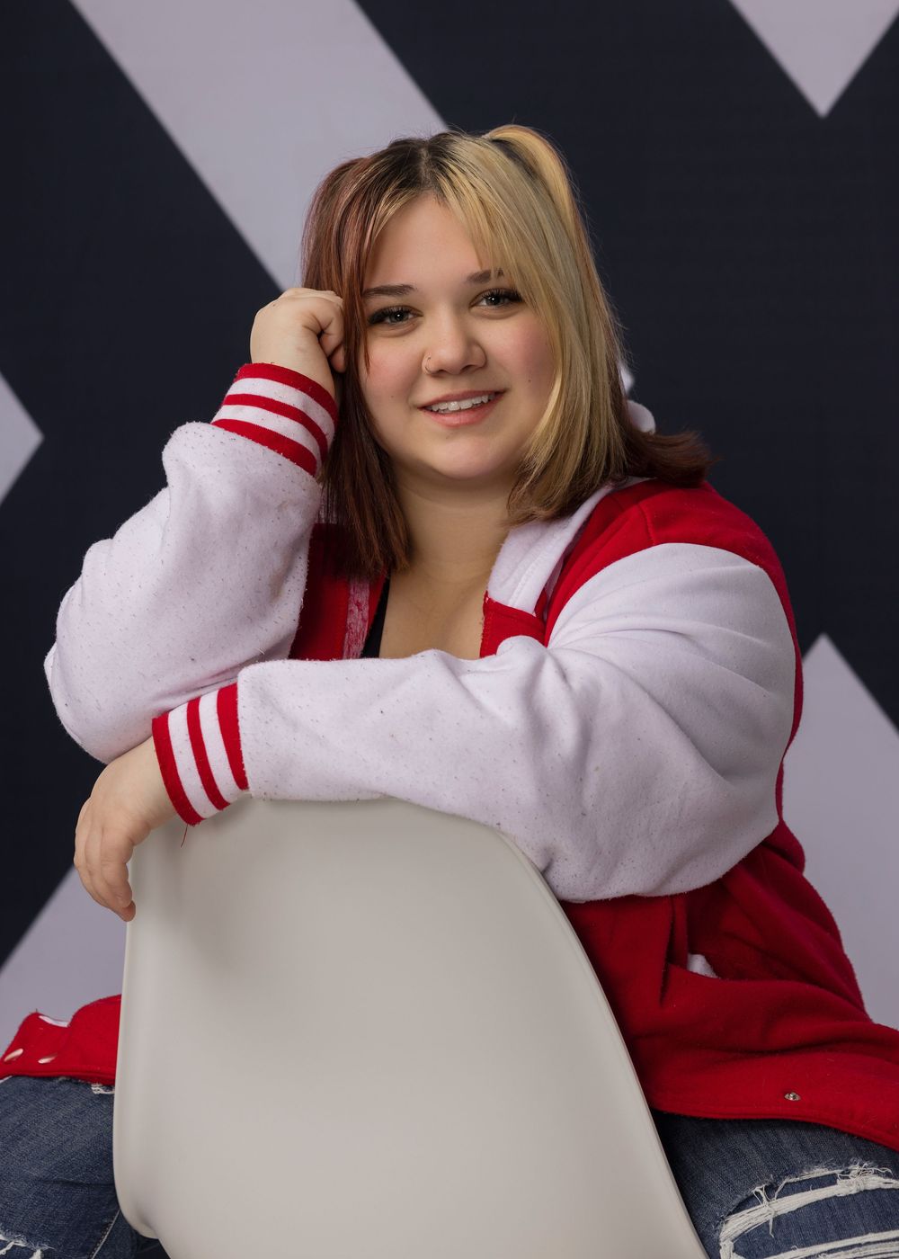 Young woman in red varsity jacket smiling, seated casually against a geometric black and white backdrop.