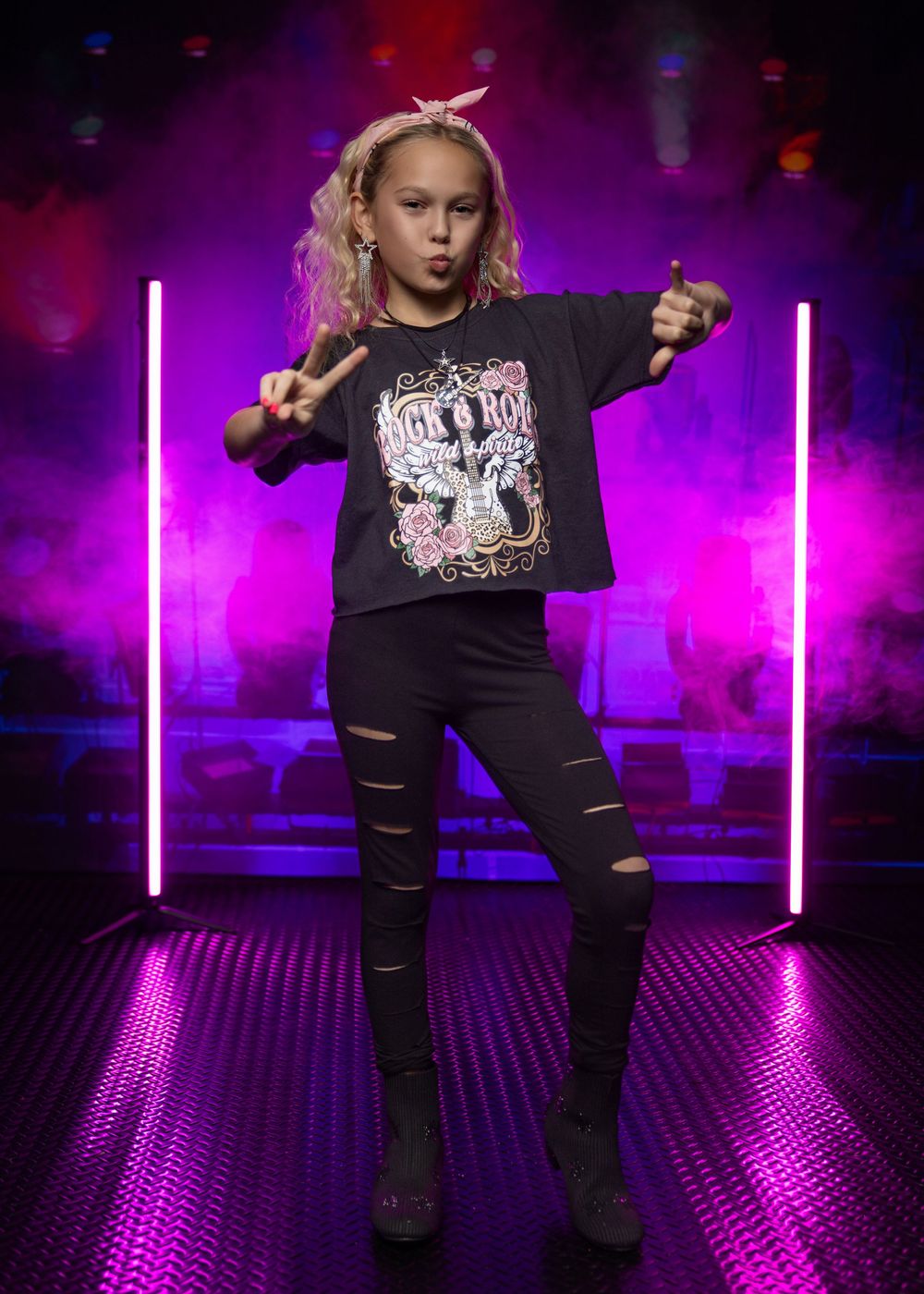 Young girl in black ripped jeans and rock t-shirt poses confidently under neon pink lights on a dark stage.