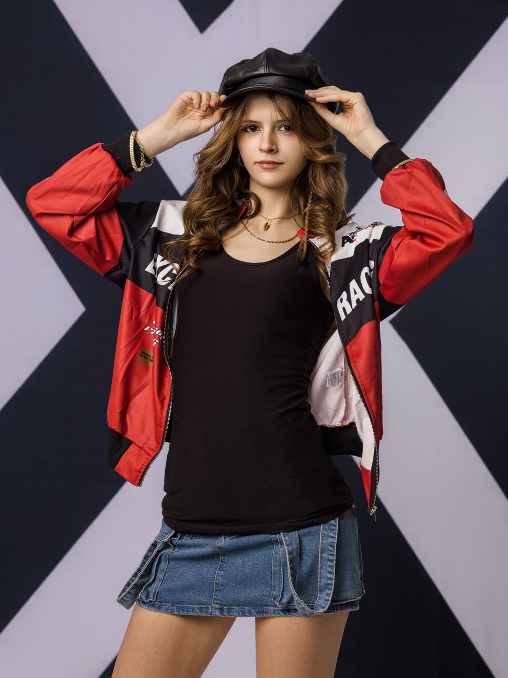Young woman in red racing jacket and denim skirt posing with black leather cap against geometric background.