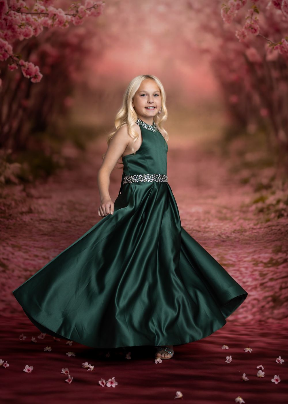 Young girl in emerald green satin gown twirling on a floral path with pink cherry blossoms in soft bokeh background.