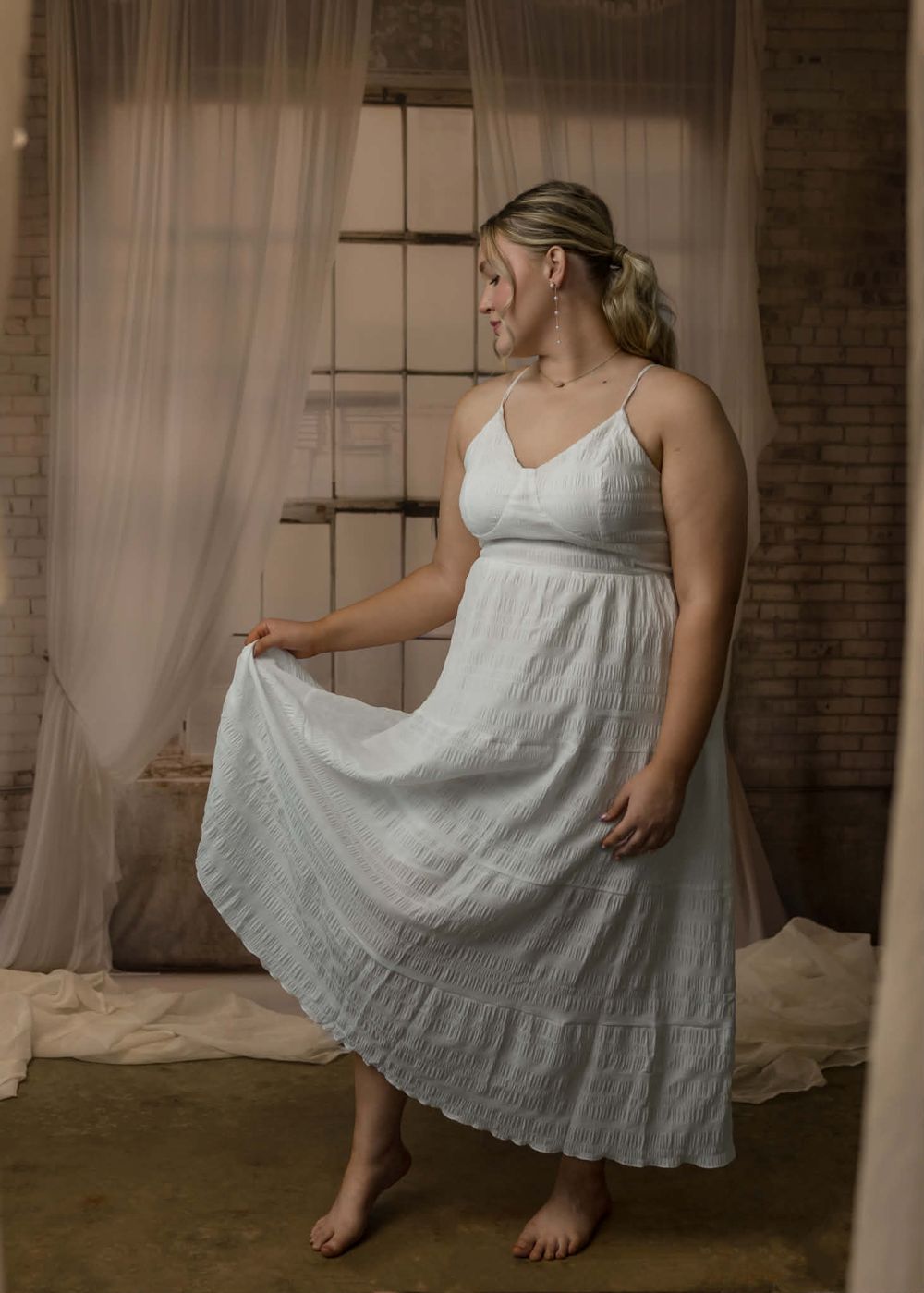 Woman in white tiered maxi dress with spaghetti straps, posing barefoot in a softly lit studio with sheer curtains.