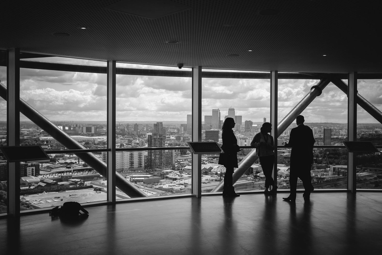 Black and white photo of silhouetted people admiring a city skyline through large windows in a high-rise building.