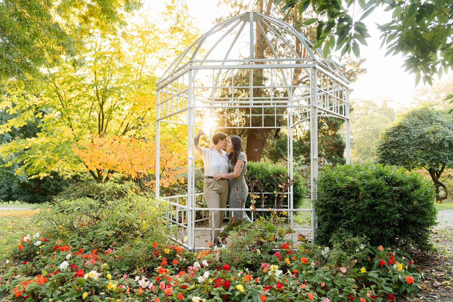 Couple sharing a romantic kiss inside a white garden gazebo surrounded by colorful flowers and lush green trees.