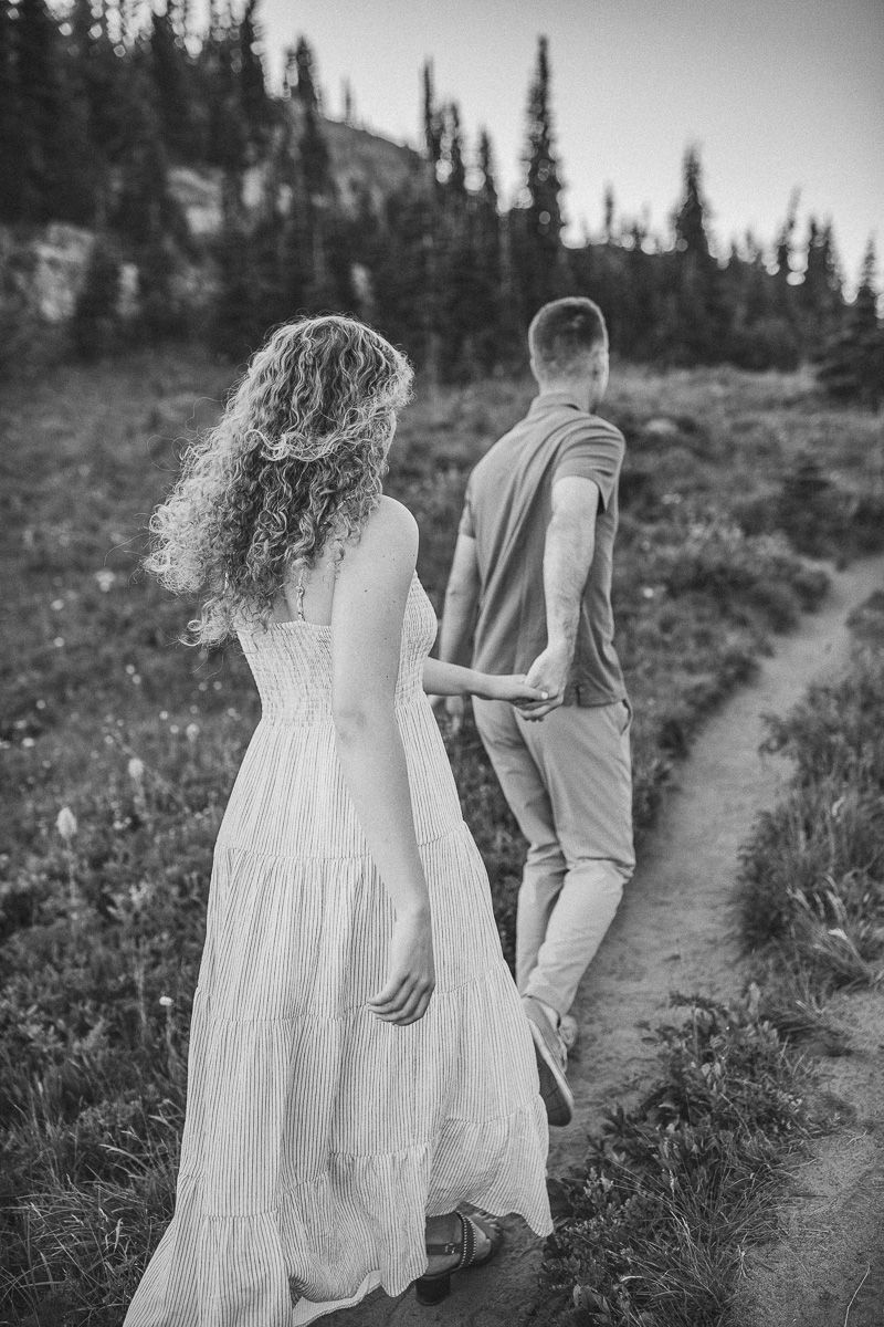Black and white photo of a couple holding hands walking on a mountain trail, woman in white dress, surrounded by pine trees.