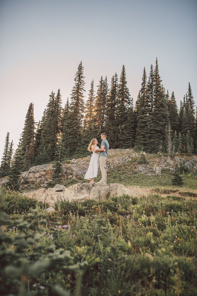 Couple embracing on rocky hilltop surrounded by tall evergreen trees at golden hour in a mountain landscape.