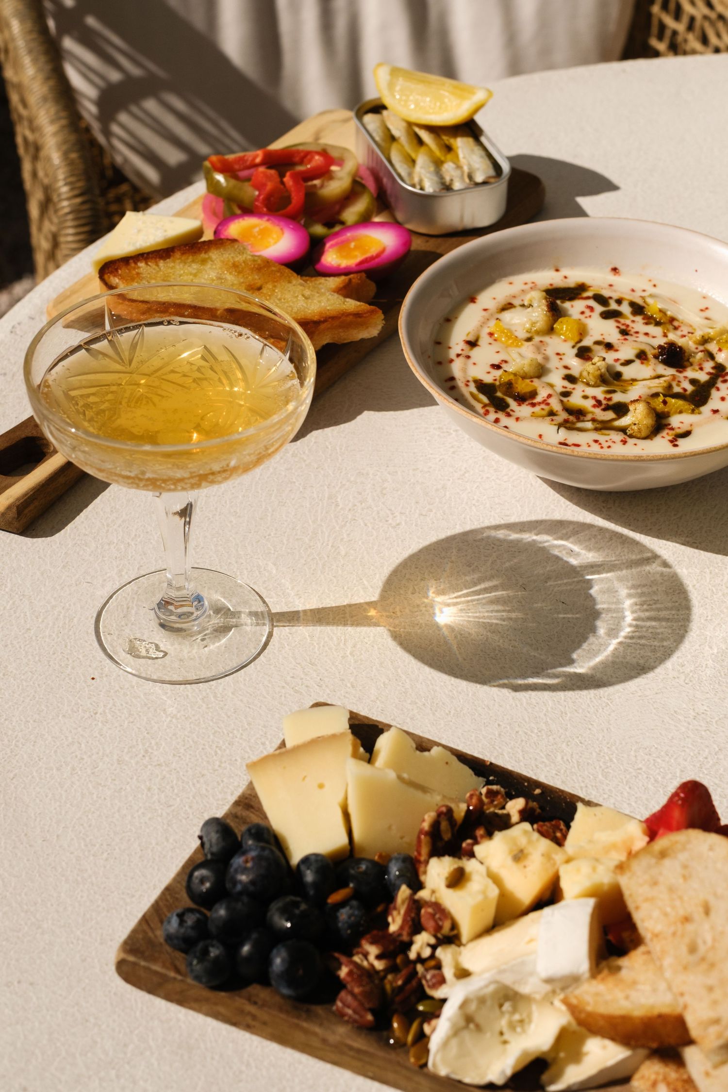 Elegant spread with a cocktail, cheese board with blueberries, hummus bowl, and appetizers on a sunlit white table.