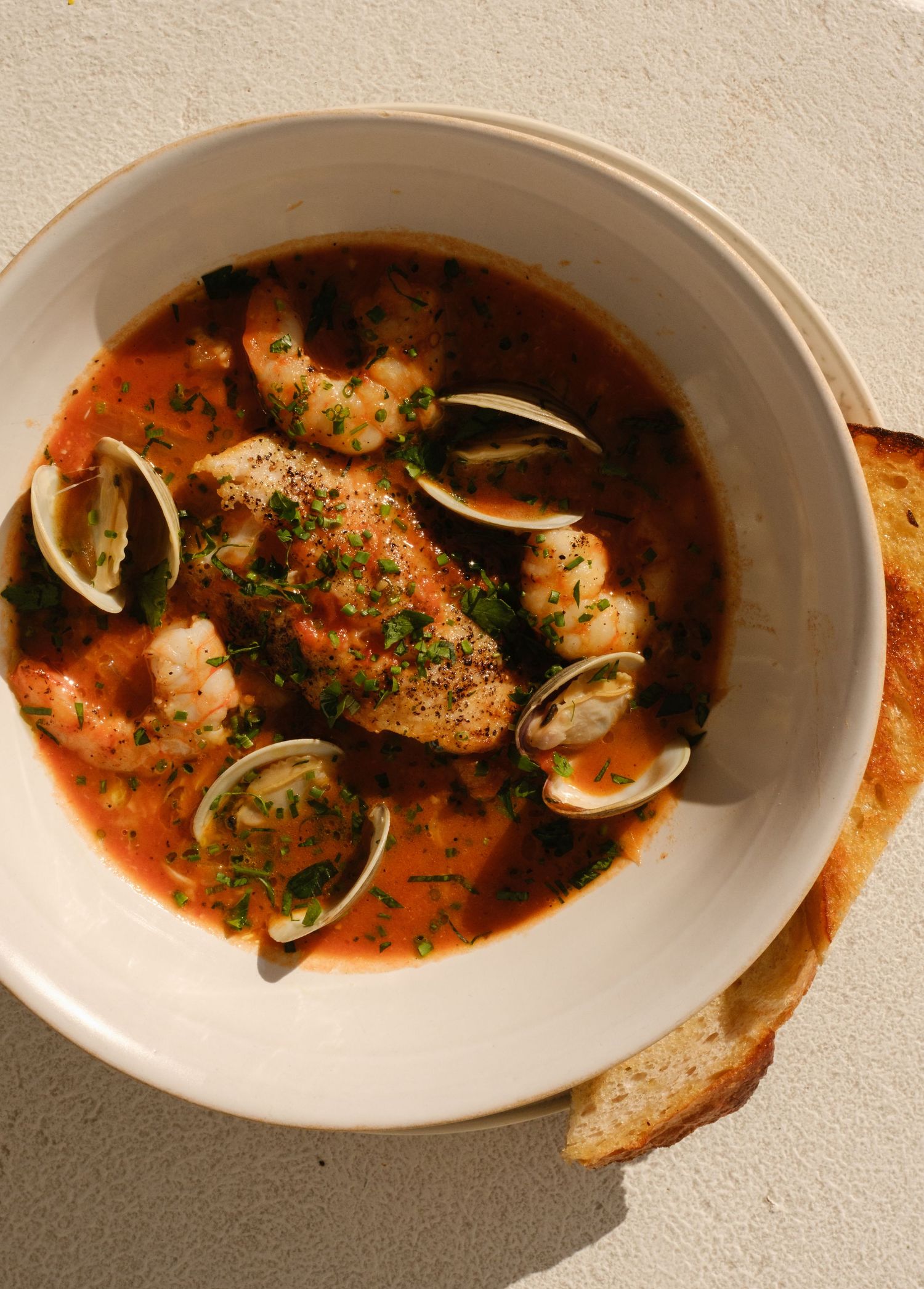 A bowl of cioppino seafood stew with shrimp, clams, and fish in tomato broth, garnished with parsley, served with crusty bread.