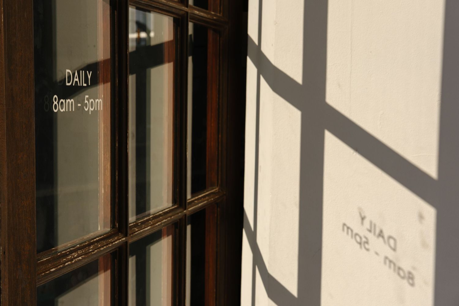 Wooden window with daily hours 8am-5pm sign casting shadow on sunlit wall.