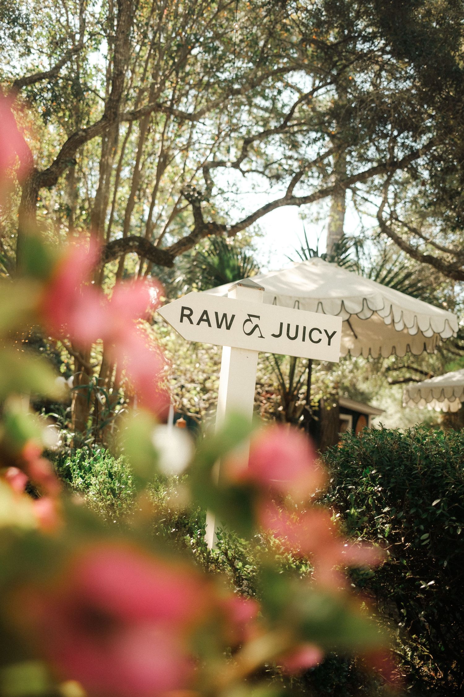 A white directional sign reading Raw and Juicy surrounded by lush greenery and pink flowers in a sunlit garden.