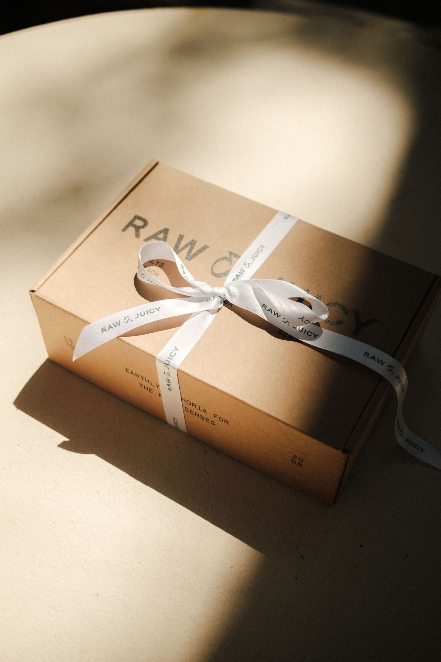 Raw Juicy branded kraft gift box tied with a white ribbon bow, placed on a neutral surface in natural light.