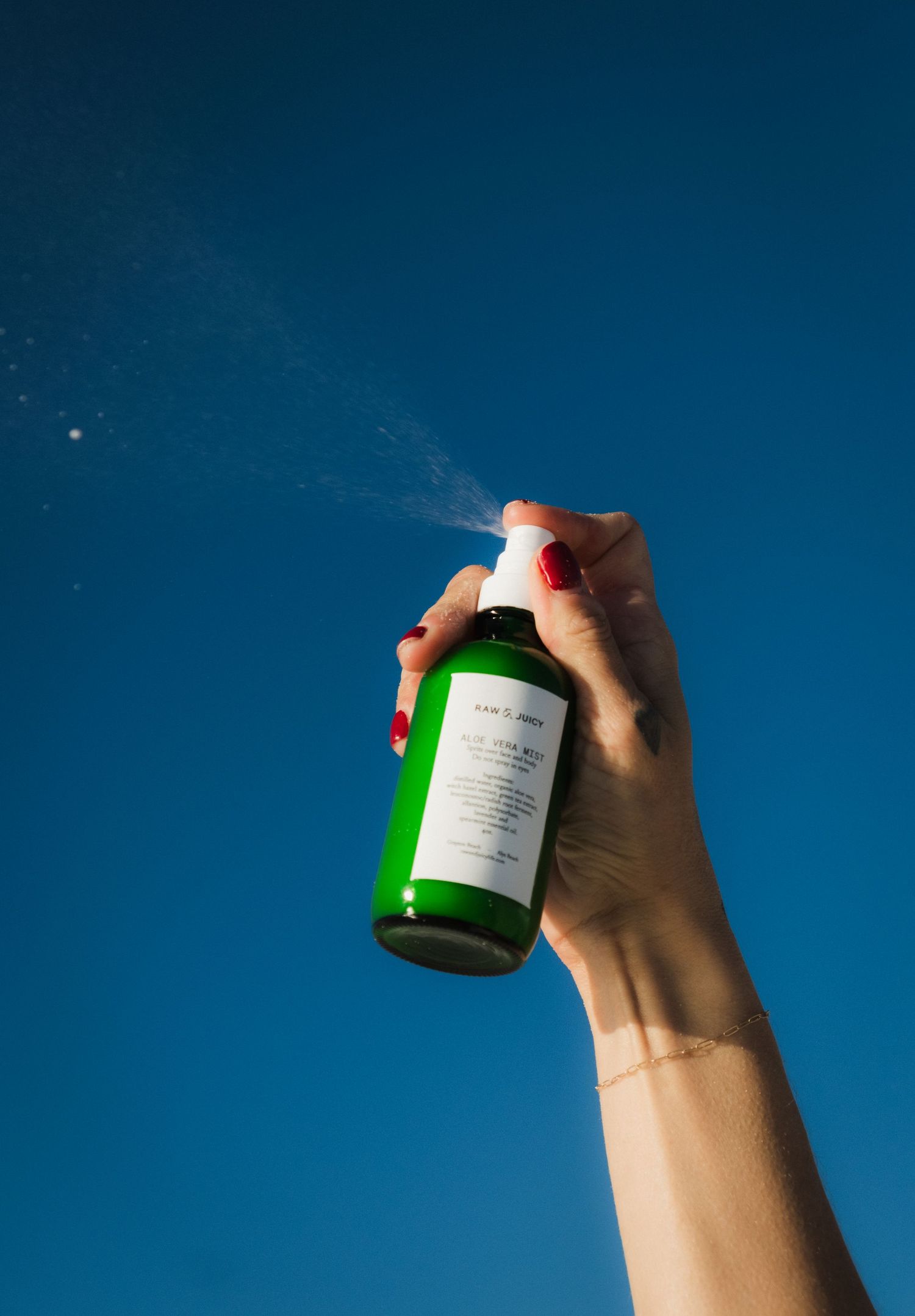 A hand with red nails sprays a green bottle of face mist against a vivid blue sky.