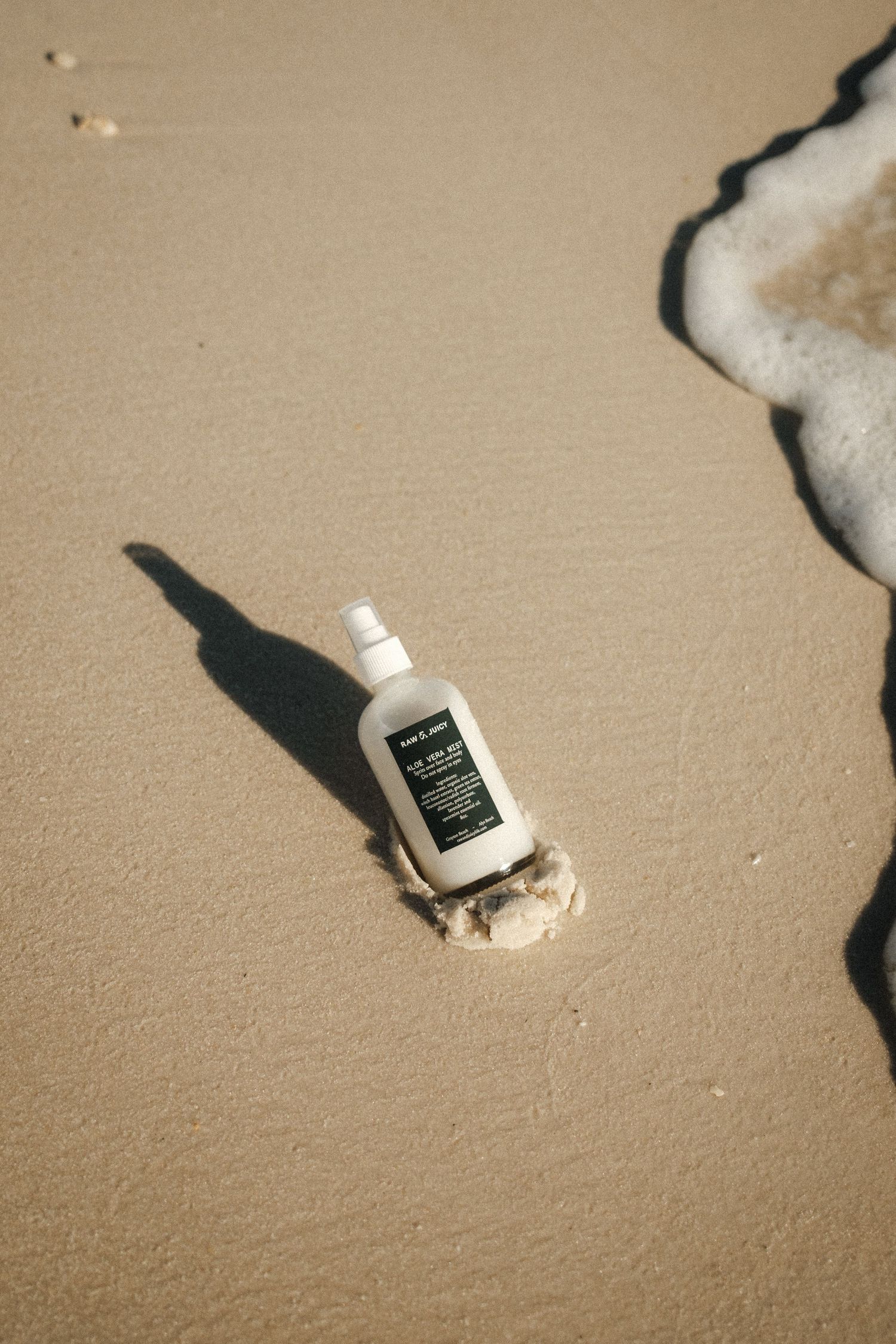 White spray bottle with dark label resting on sandy beach near ocean waves and small rocks.